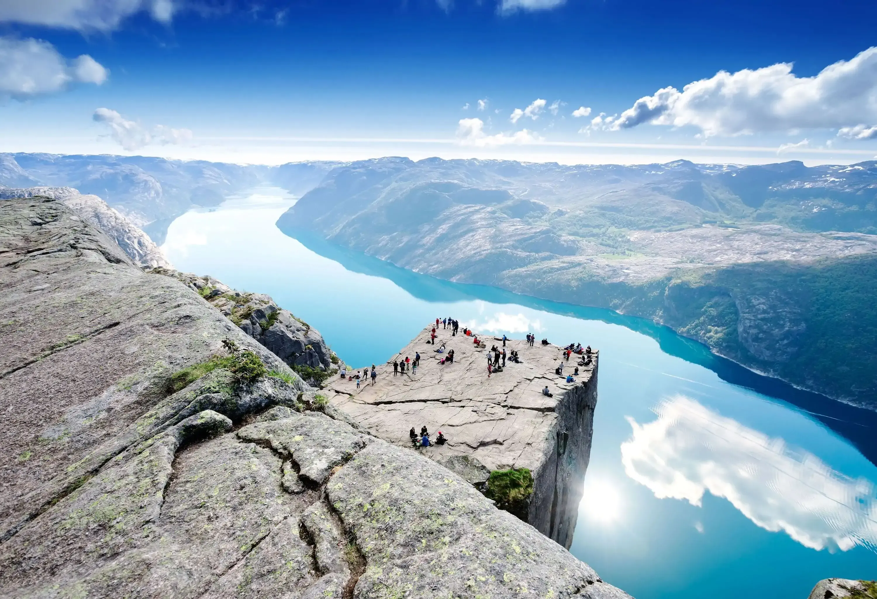 A group of hikers milling around the flat surface of a cliff, with an admirable view of a fjord that reflects the sun and the clouds.