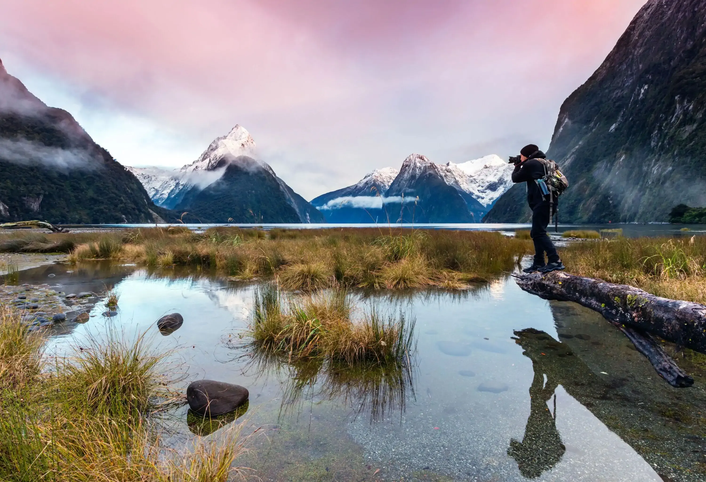 A male photographer standing on a log across a marsh to snap the stunning snow-capped mountains.