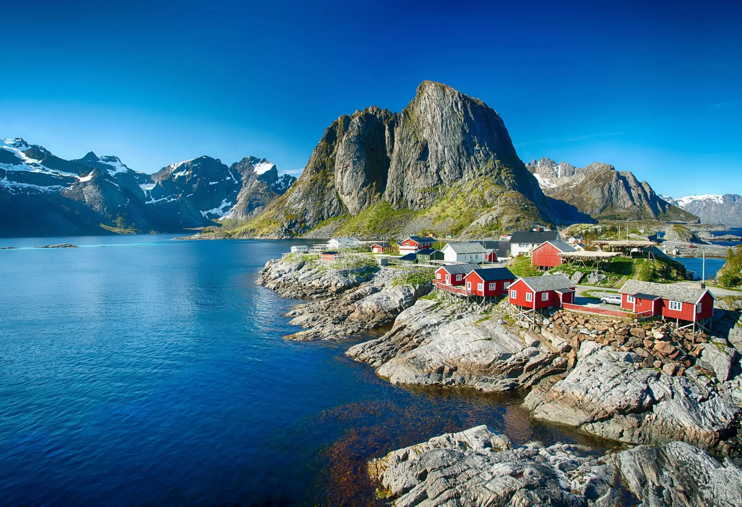 Red wooden houses gracefully perch on a rocky bank, framed by snow-dusted, rugged mountains.