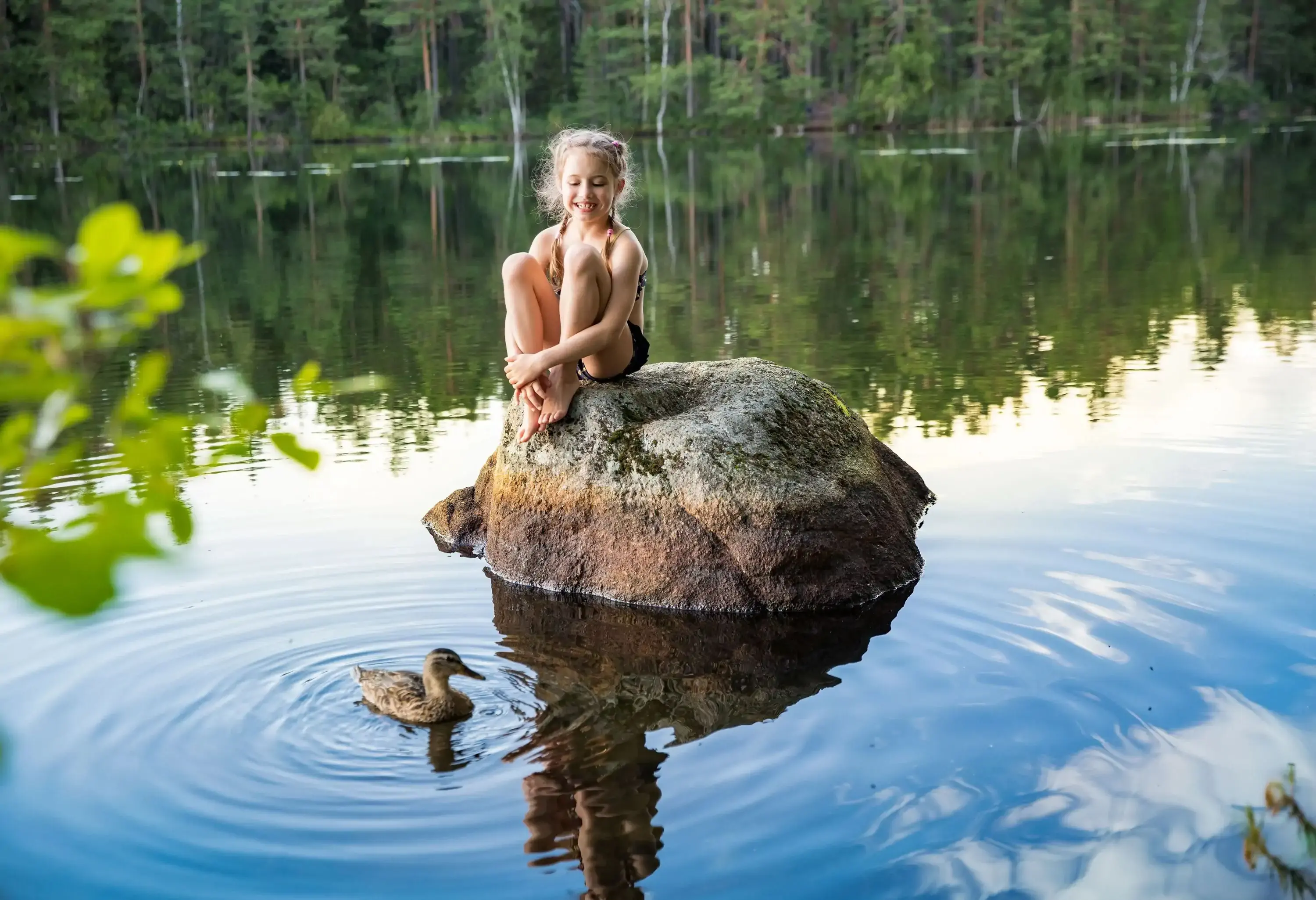 A little girl sitting on a rock in a lake and watching a duck swim in the water.