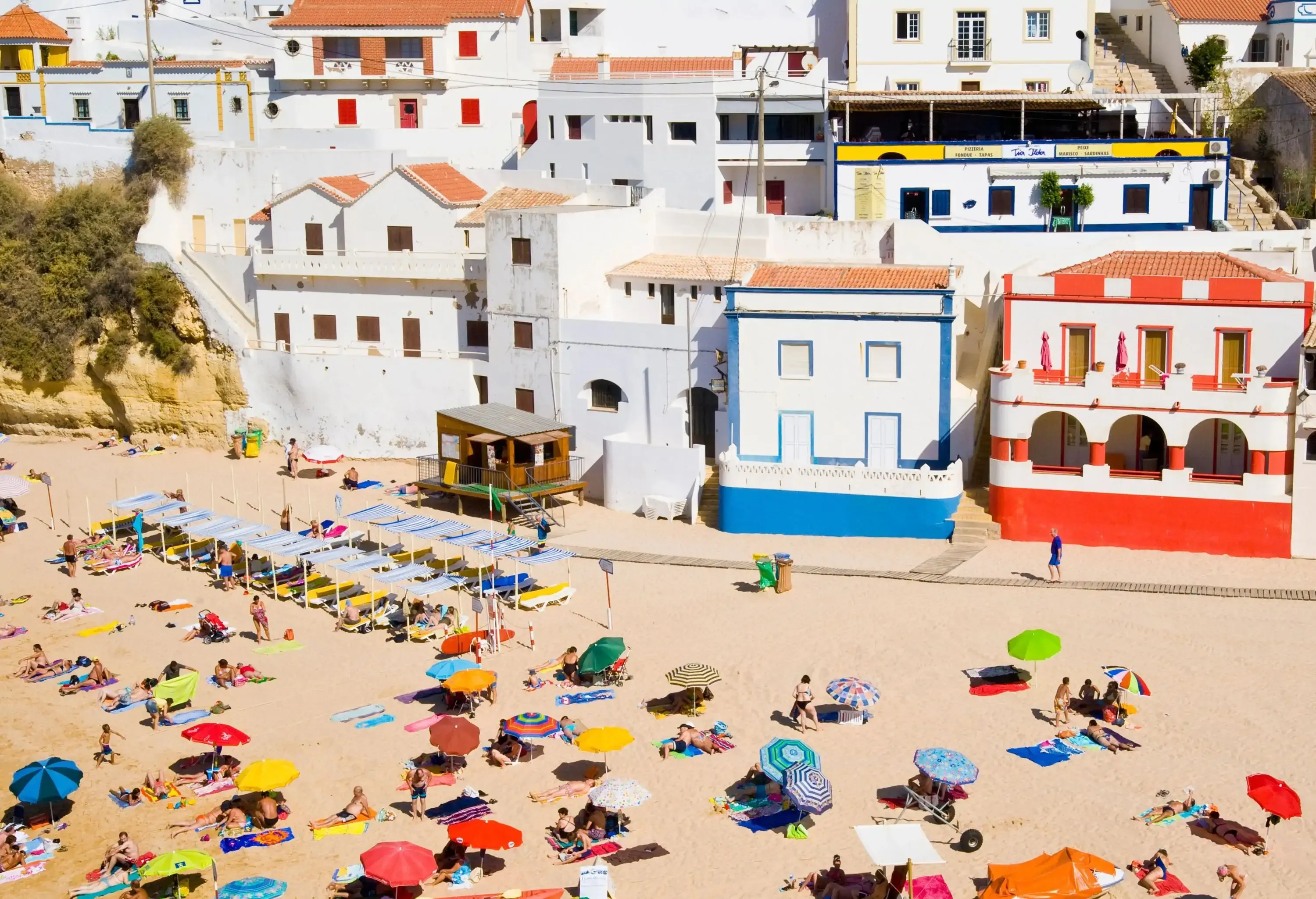 People sunbathe on the beach with colourful parasols among colourful resort buildings uphill.