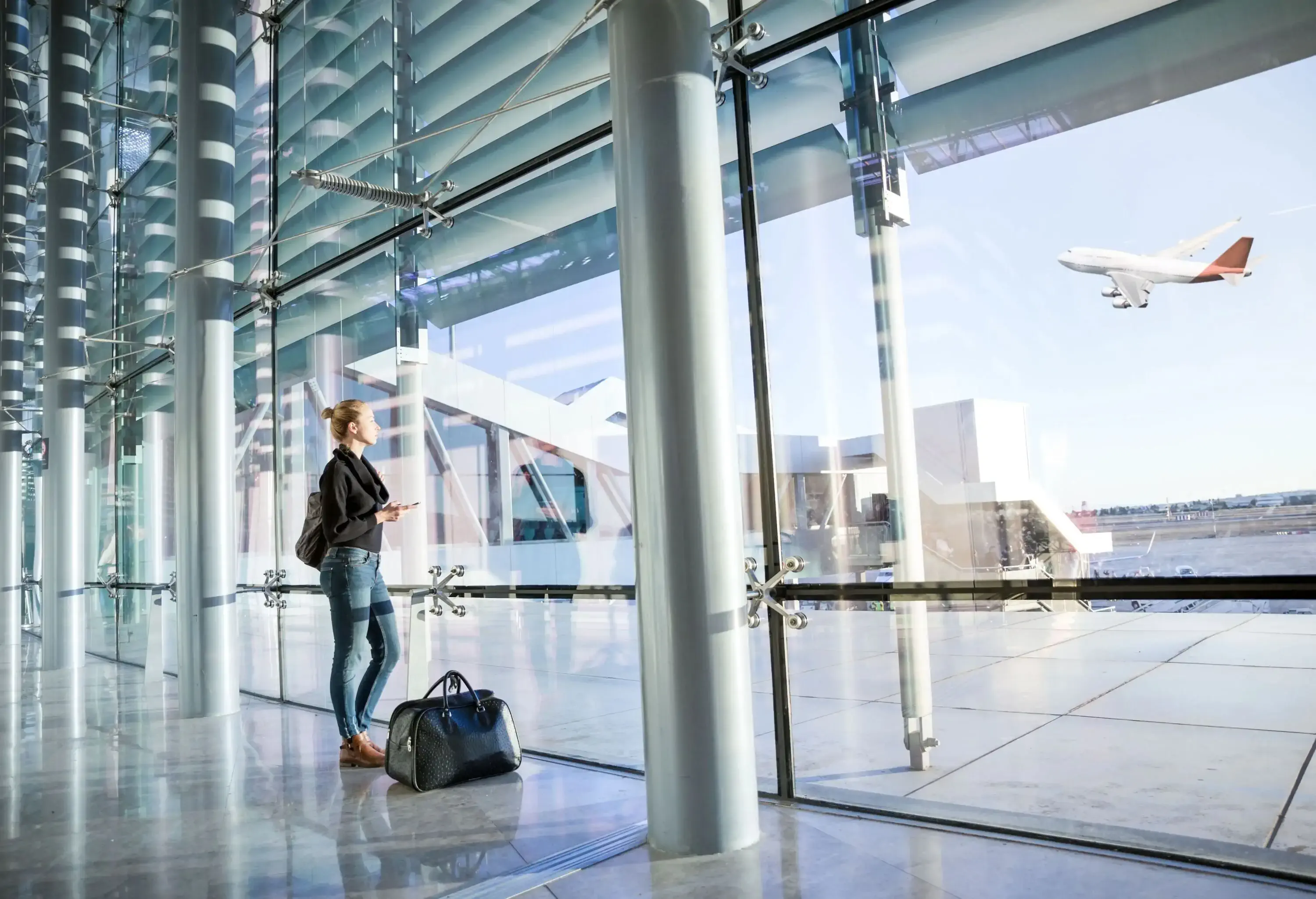 Young casual female traveler at airport, holding smart phone device, looking through the airport gate windows at planes on airport runway.