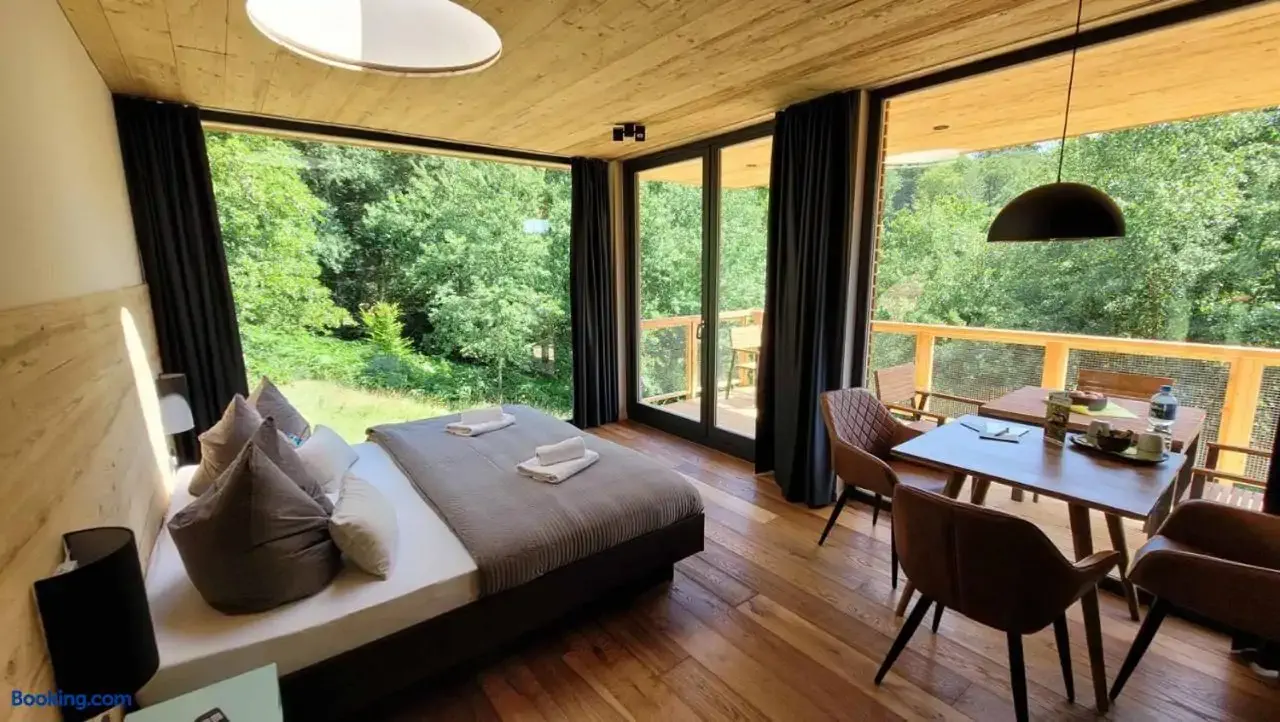 A cozy, modern wooden cabin interior with large windows overlooking lush greenery, featuring a bed, dining area, and minimalist decor.