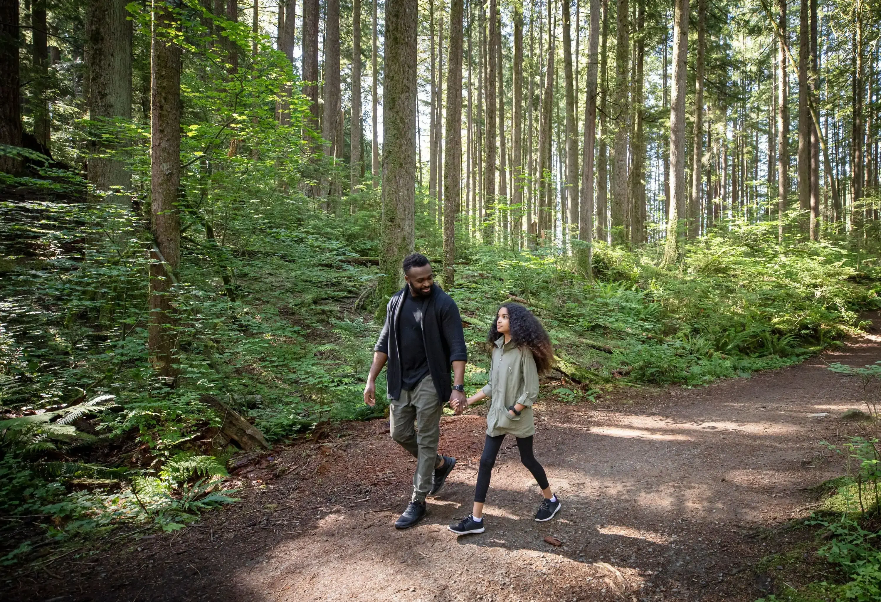 A man holds the hand of a girl as they walk on a trail in a forest with tall trees.