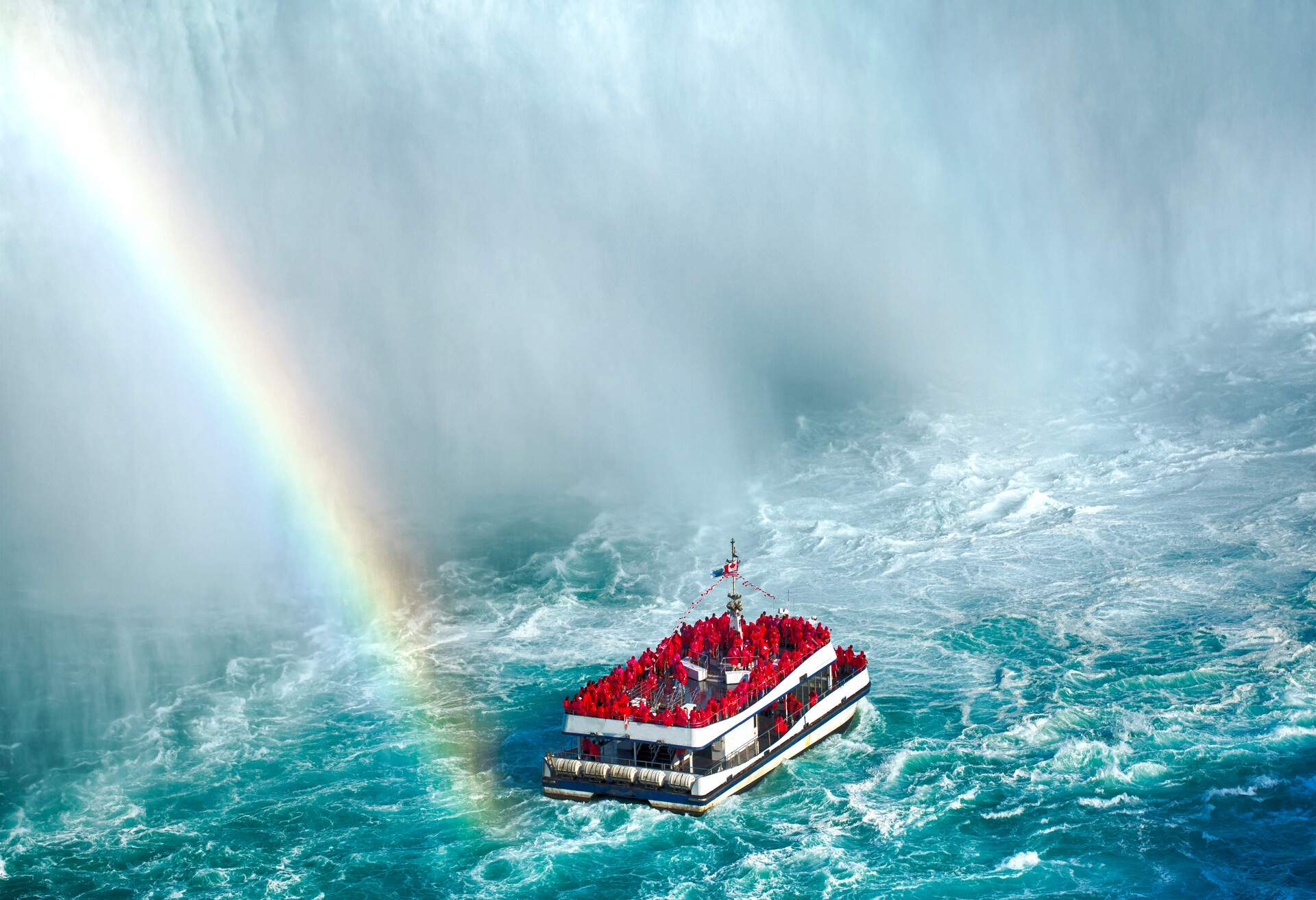 Passengers donning red plastic coats relish a thrilling boat ride at the base of Niagara Falls, with a radiant rainbow embellishing the breathtaking scene.