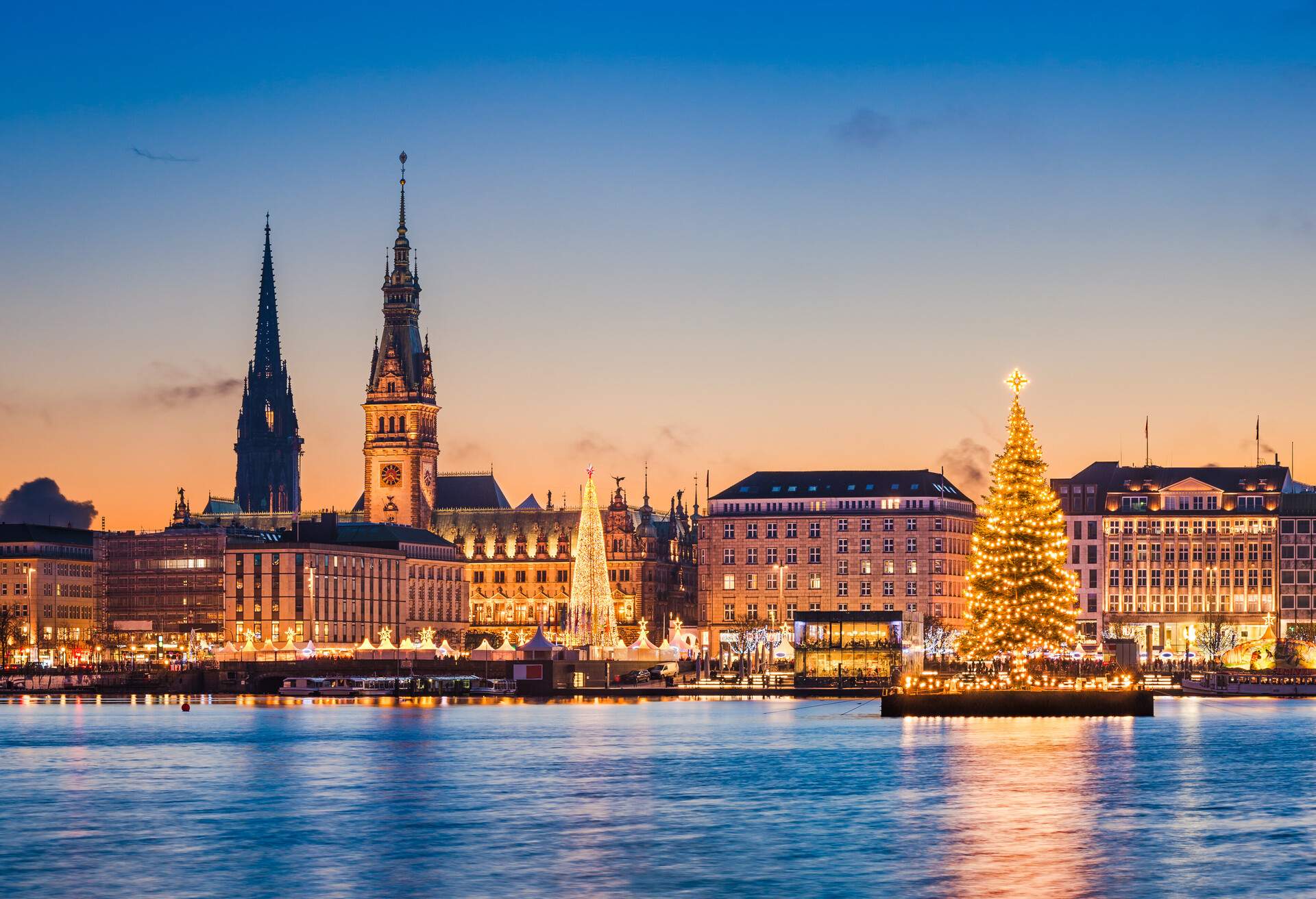 A glowing Christmas tree and decorated booths along the harbour with skyline views.