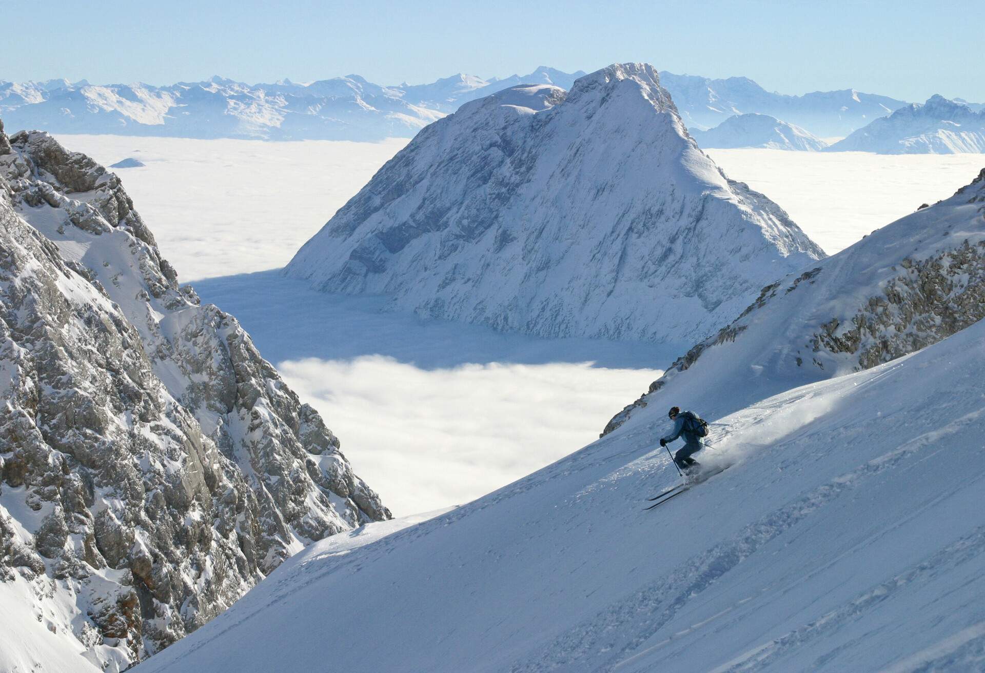 A skier descending a steep snow slope with views of rocky mountains covered in thick fog.
