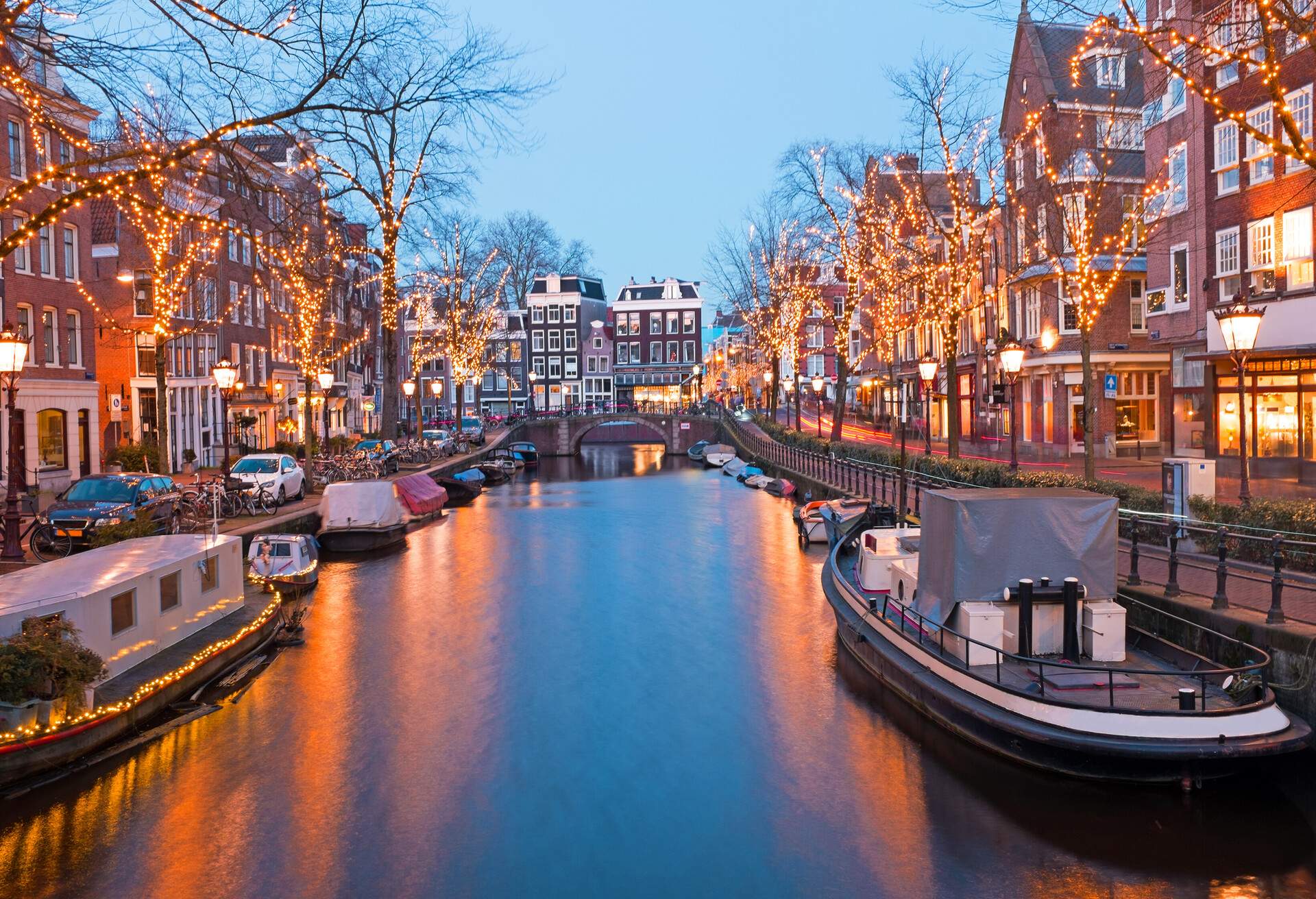 A calm canal bordered by docked houseboats, light-wrapped trees, and adjacent buildings.