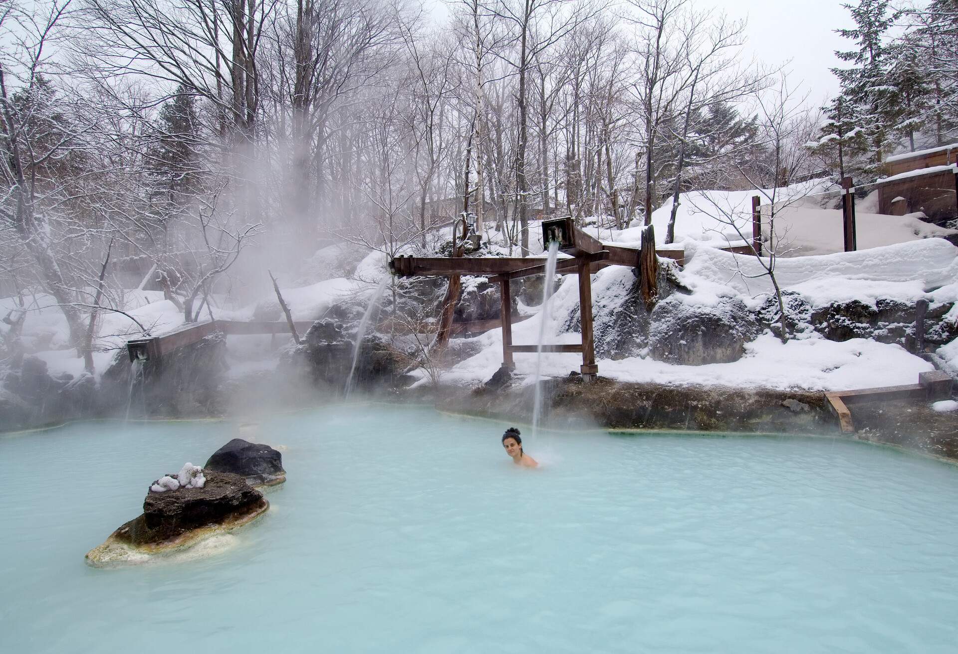 A woman dips into a steaming outdoor hot spring surrounded by frosted trees and snow-covered landscapes.