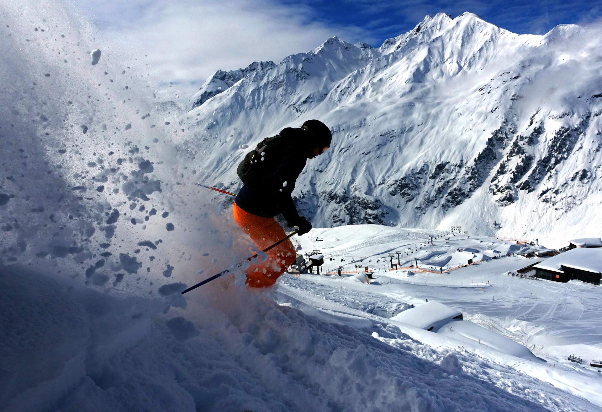 A skier slides down the snow-covered terrain with powdery snow behind and views of snow-covered mountains.