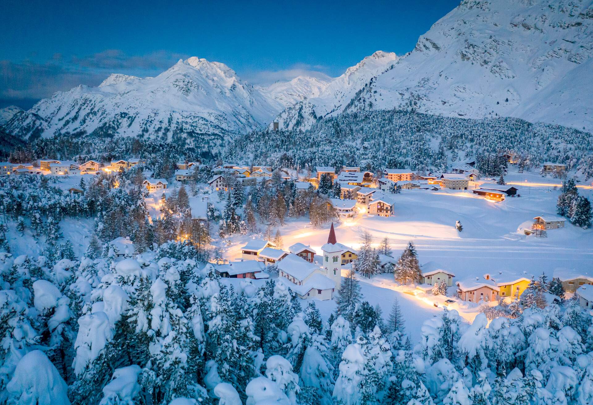 Aerial view of a brightly lit alpine village in the valley of the forested mountains covered in deep snow.