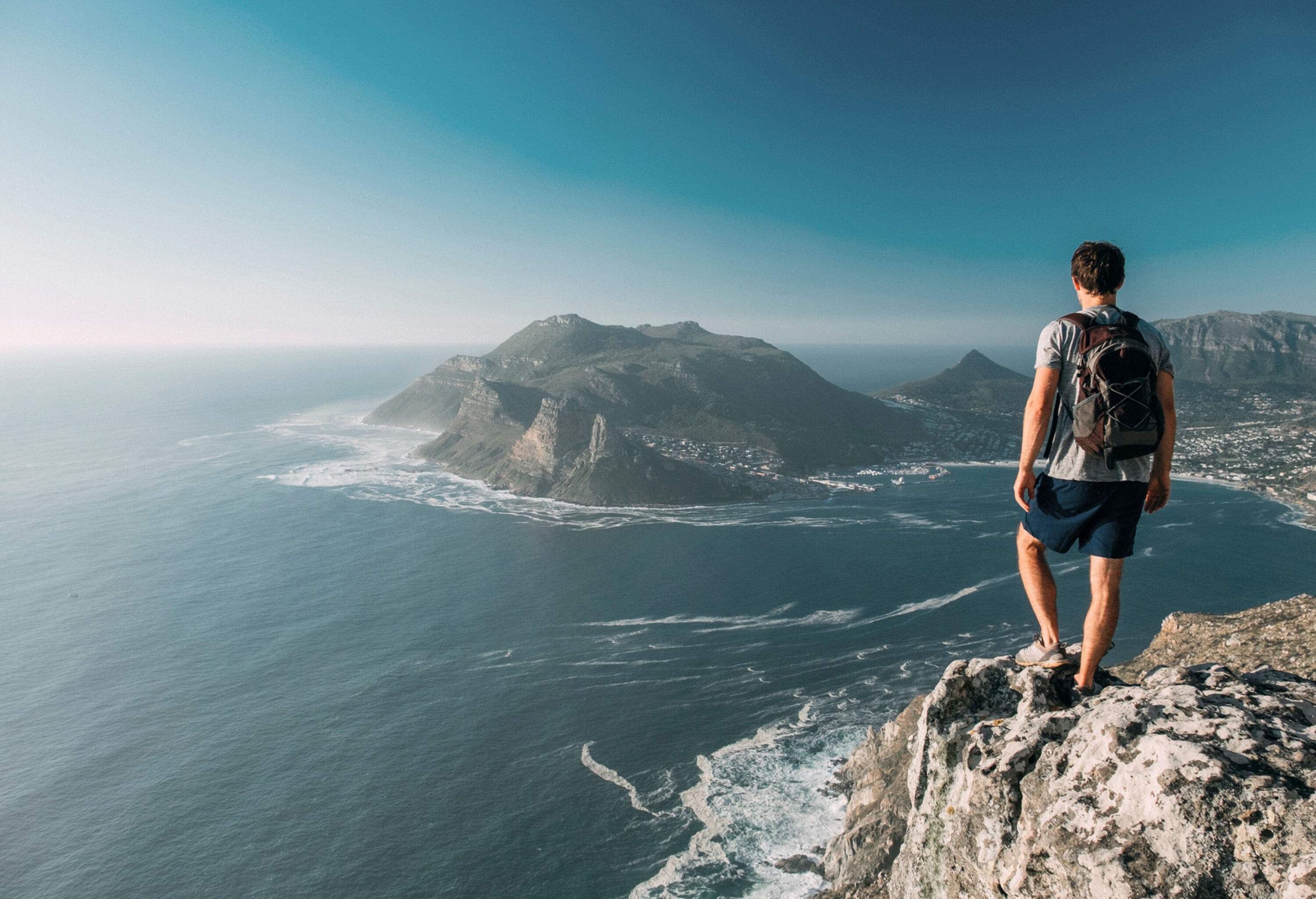 A man with a backpack stands on a rock cliff and stares at the vast ocean, coastal city, and surrounding mountains below.