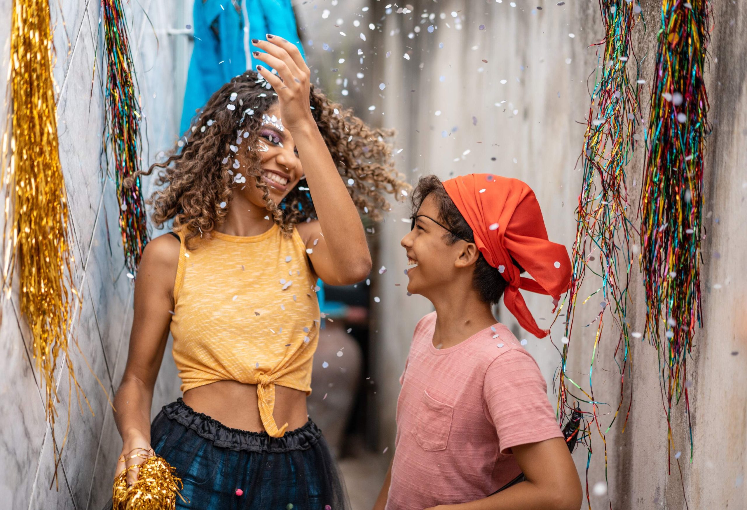 Two kids, one in orange shirt and the other in a red bandana, laugh together under the shower of confetti.