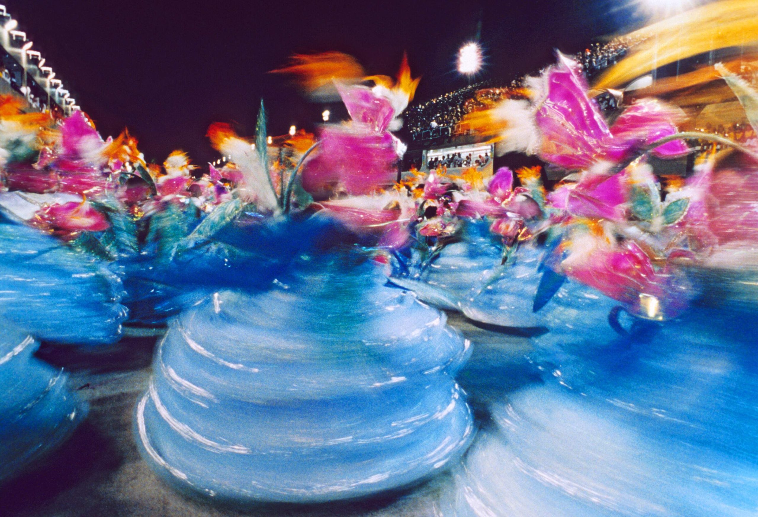 A blurry image of dancers in colourful traditional costumes dancing on the street.