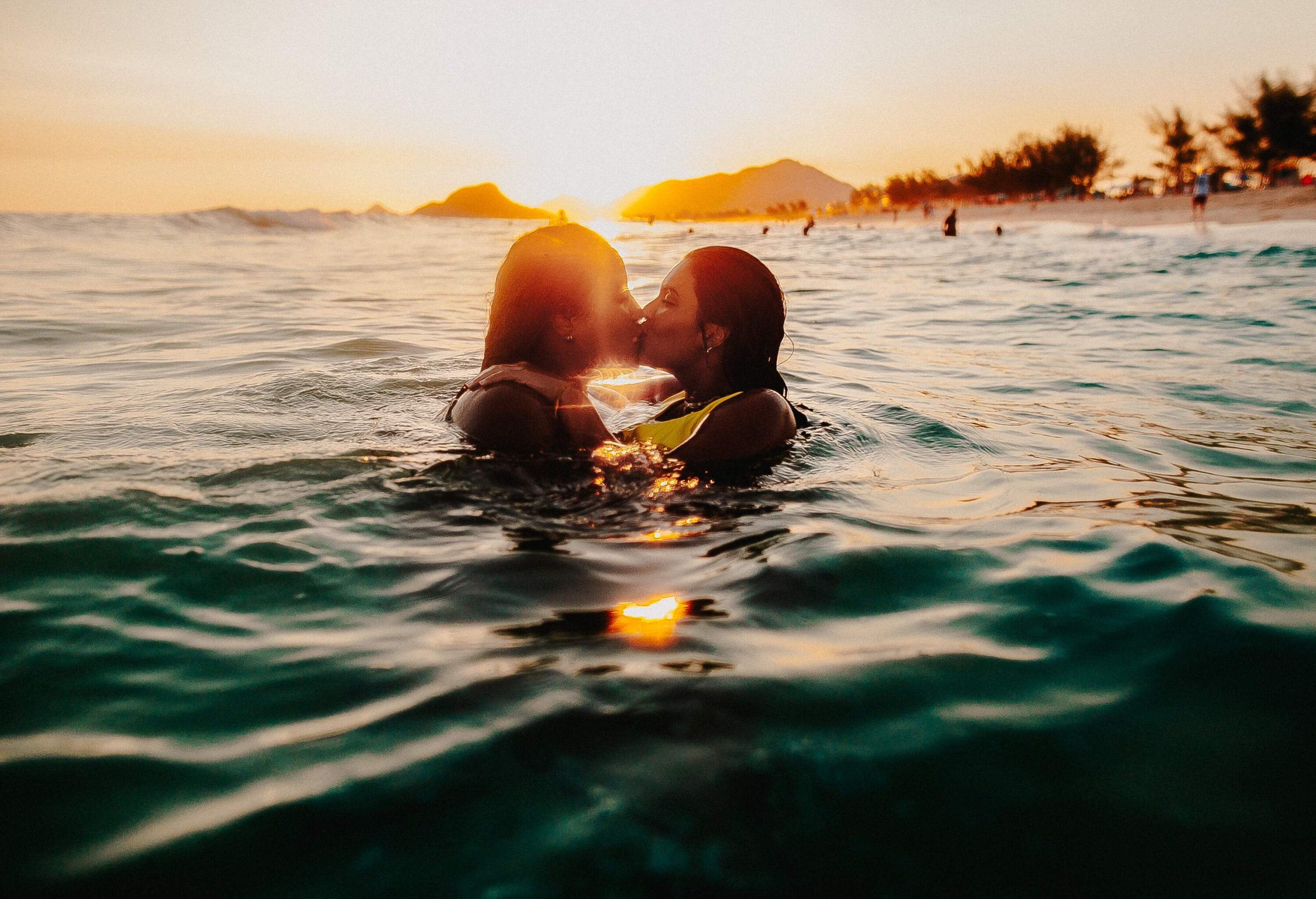 A female couple shares a kiss while immersed in sea water with the sun setting in the background.