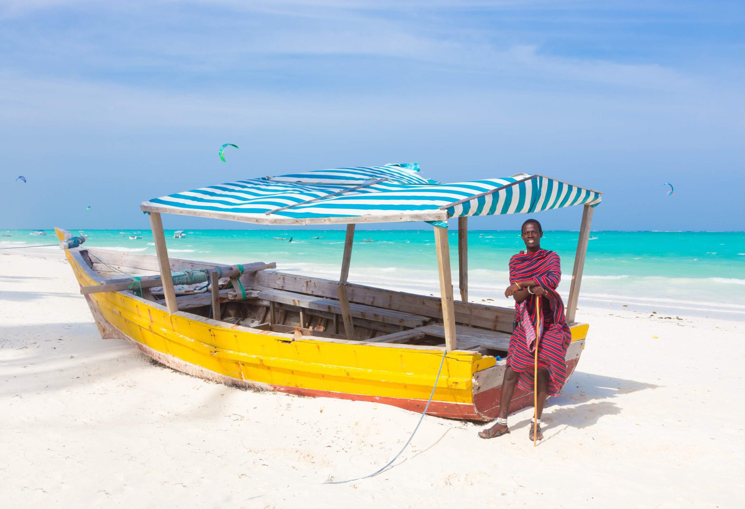 A Maasai man leans on a wooden stick, standing beside a colourful boat docked on the soft sands of the beach.