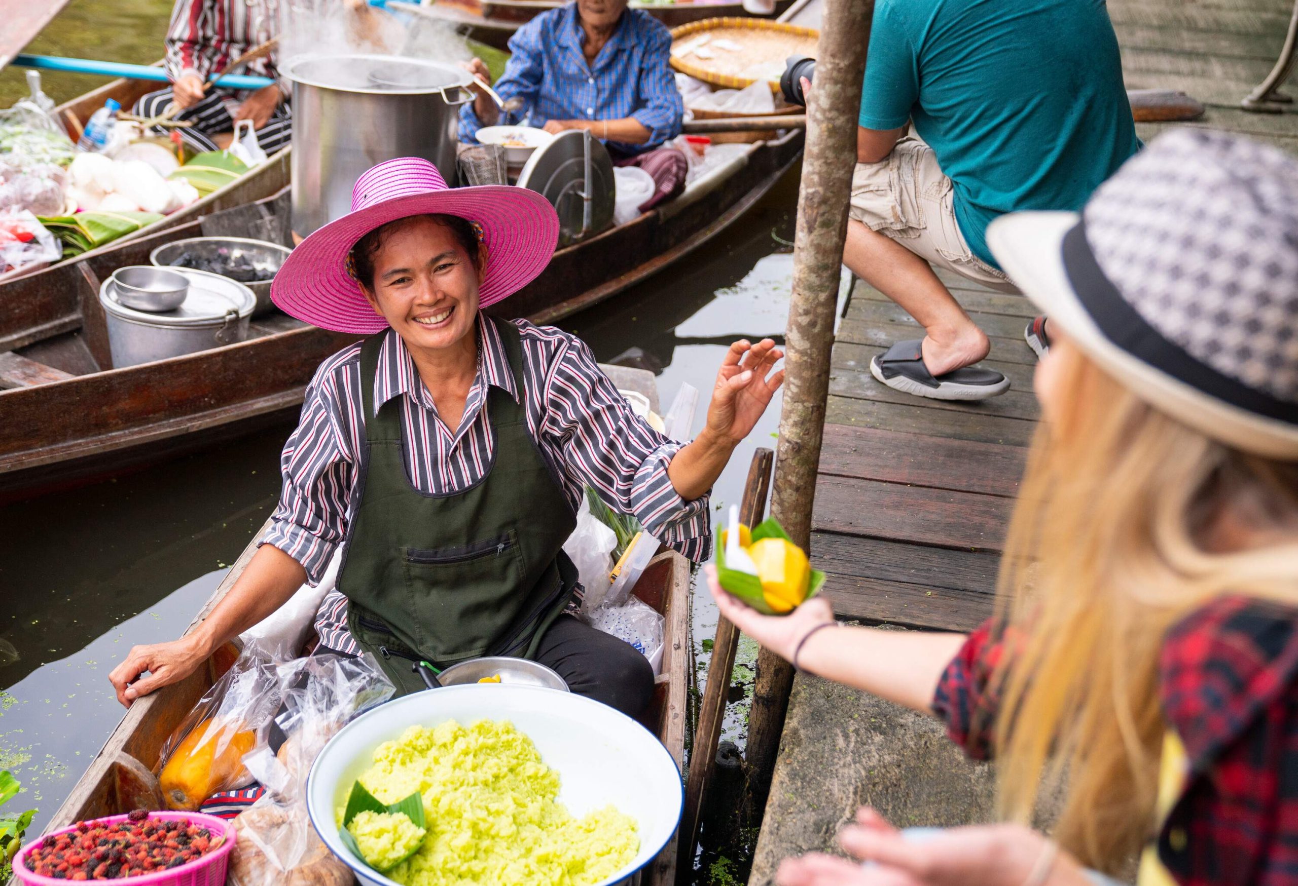A tourist extends her hand with snacks to a smiling vendor wearing a wide-brimmed hat on a boat in a floating market.