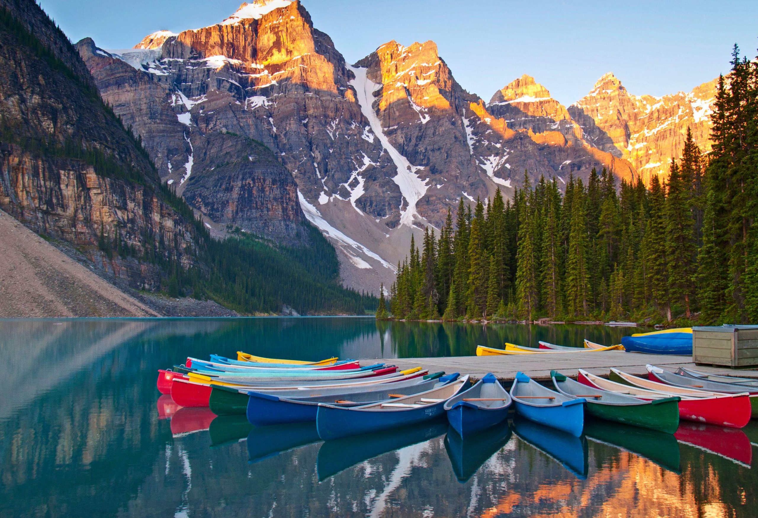 Colourful canoes surrounding a wooden dock on a lake nestled between snow-capped mountains and trees.