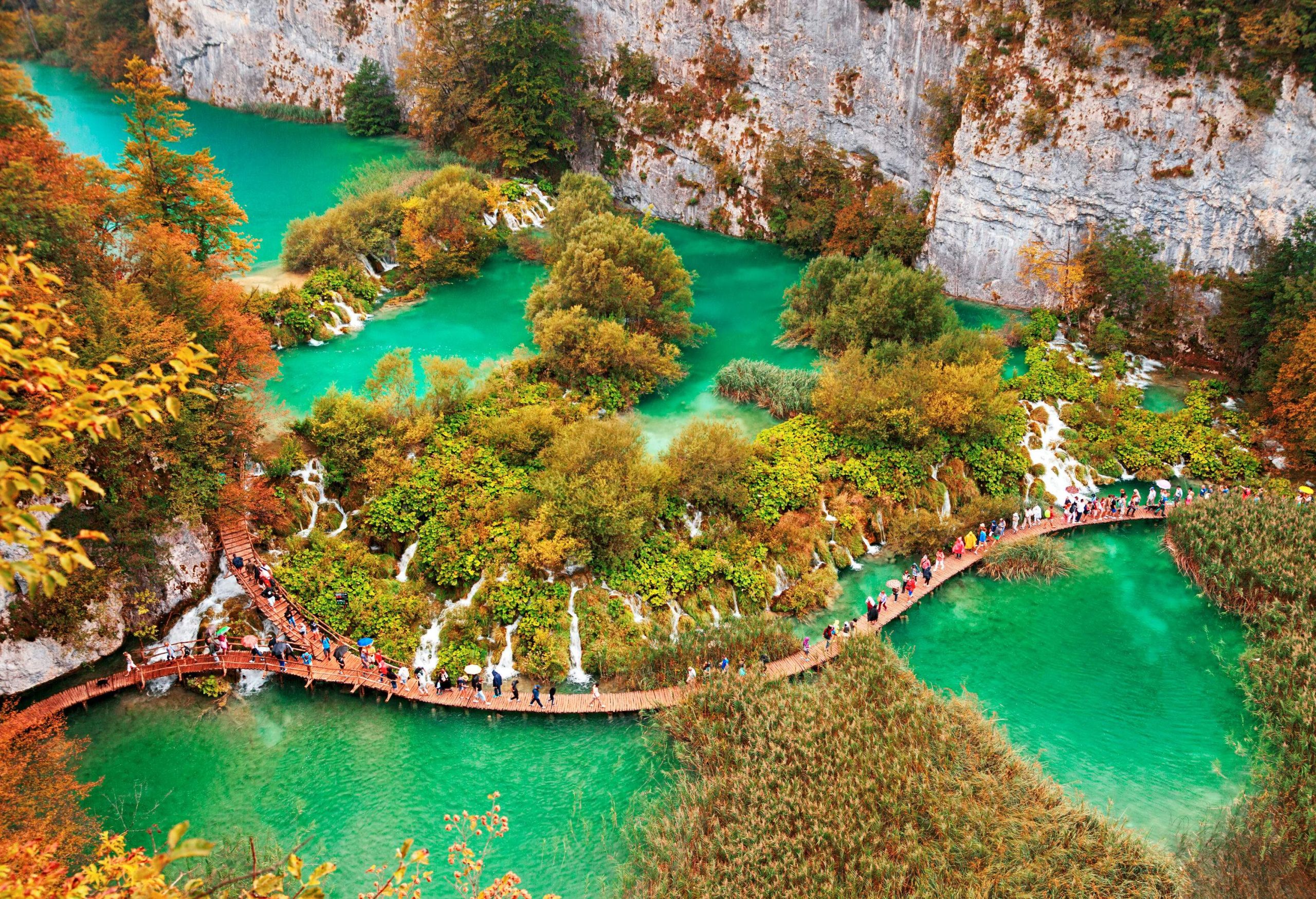 A boardwalk with people passing through suspended over a green lake surrounded by autumn trees along the side of a rocky mountain.