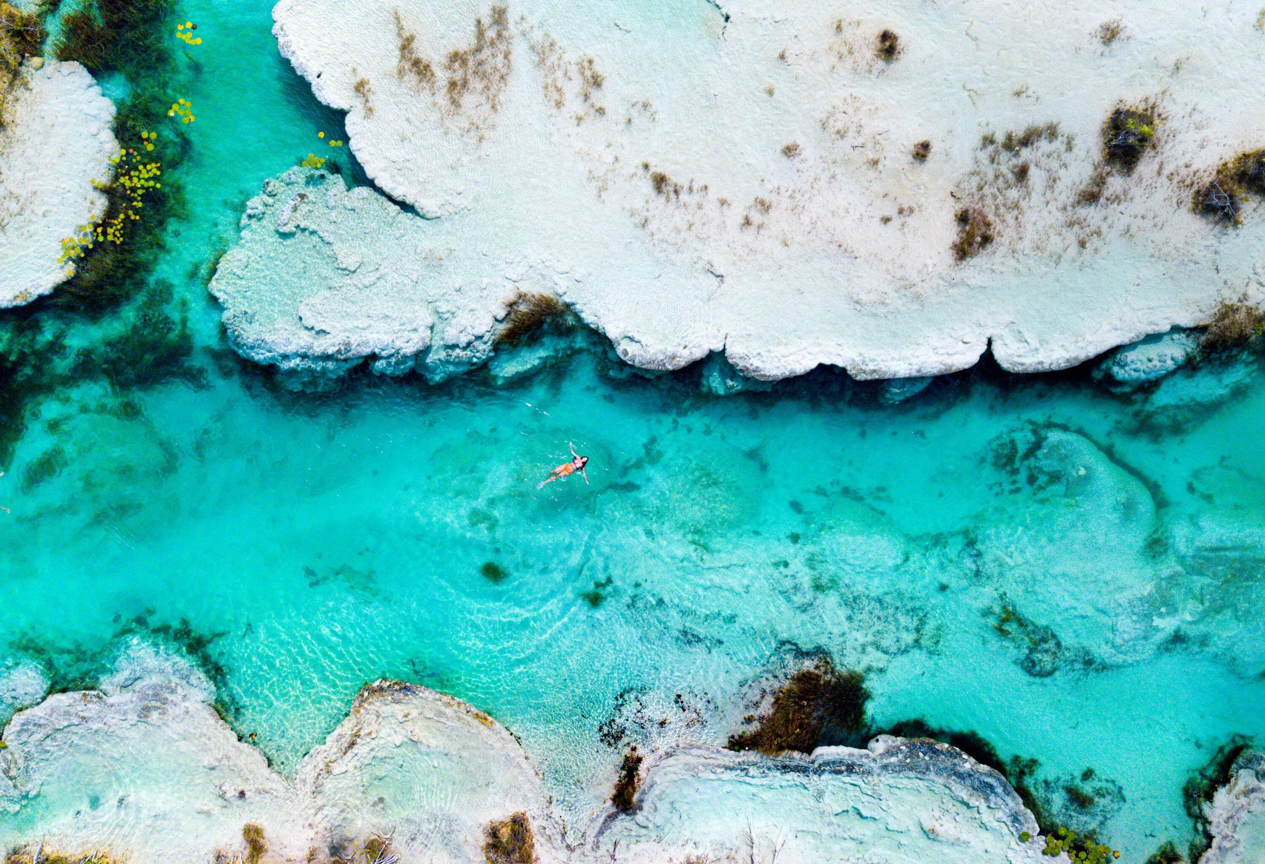 A woman in a bikini floats on the crystal-clear turquoise water in a narrow lagoon enclosed by layered rocks.