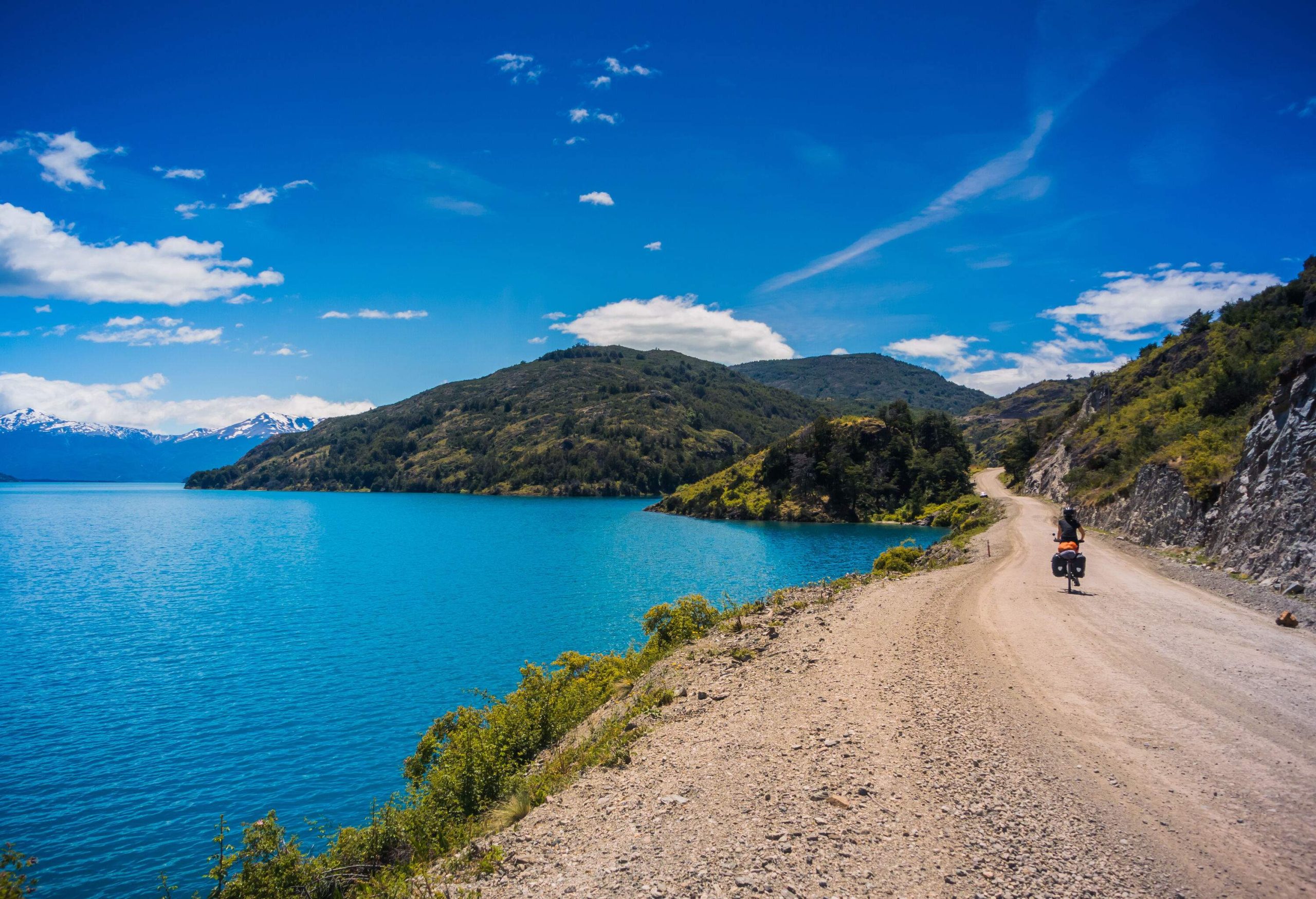 A person bicycling on a sandy sea coastal road with a view of forested mountains and snowy mountains against a clear blue sky.