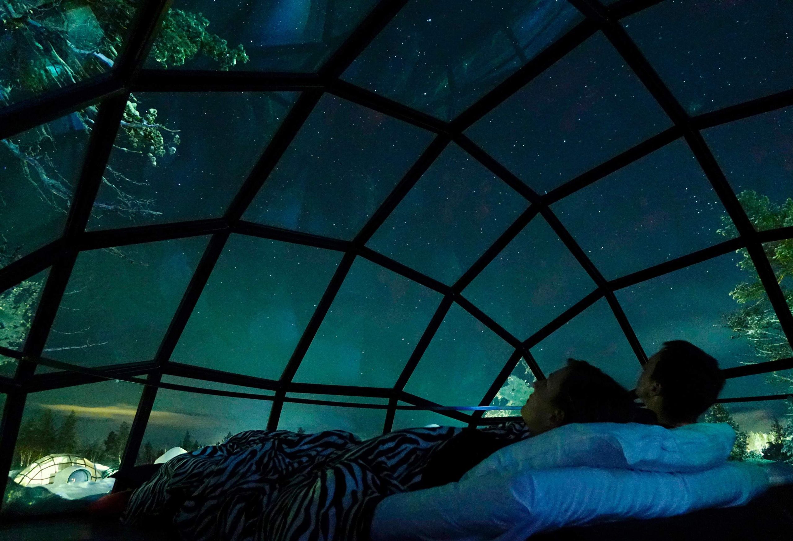 A couple lies on a bed in a glass igloo looking at the stars in the sky.