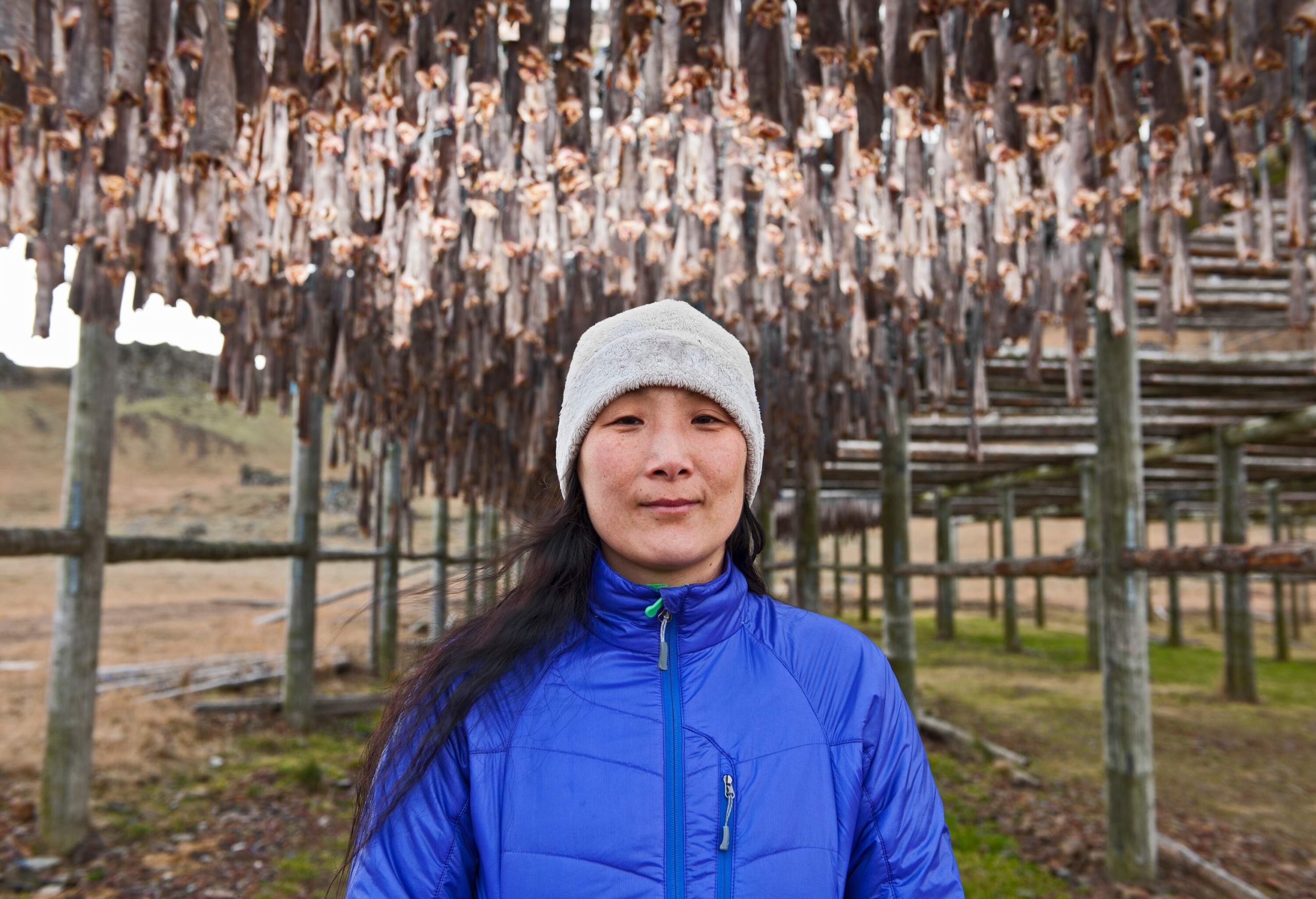 A woman in a blue jacket standing beneath rows of fish hanging on wooden racks to dry in the air.
