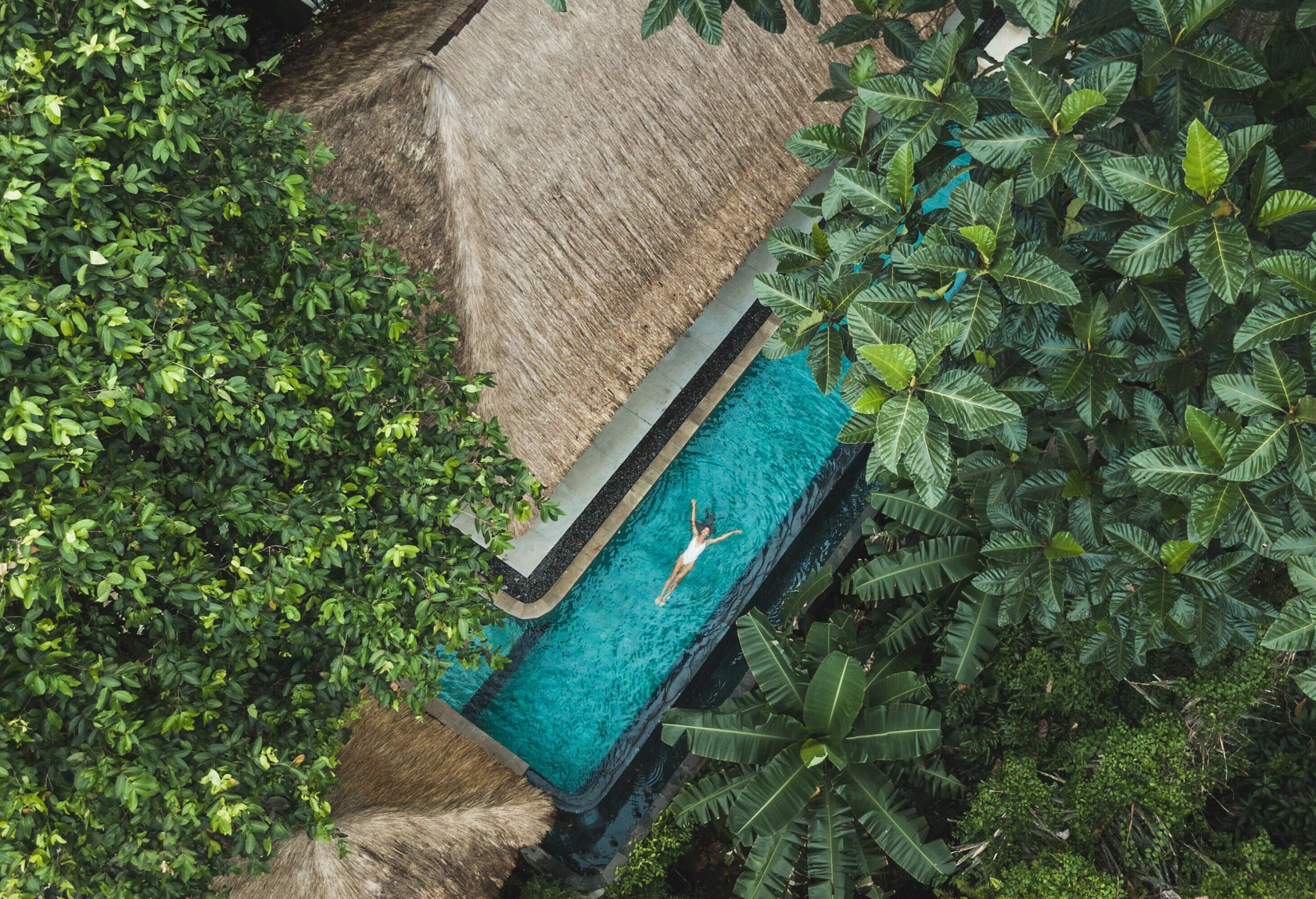 A woman in a white swimsuit floats in a pool next to a straw-roofed jungle cottage.