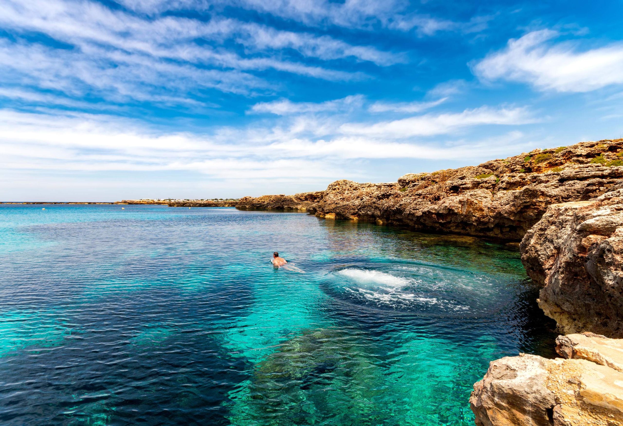 A man swimming in the blue ocean surrounded by low rock cliffs.