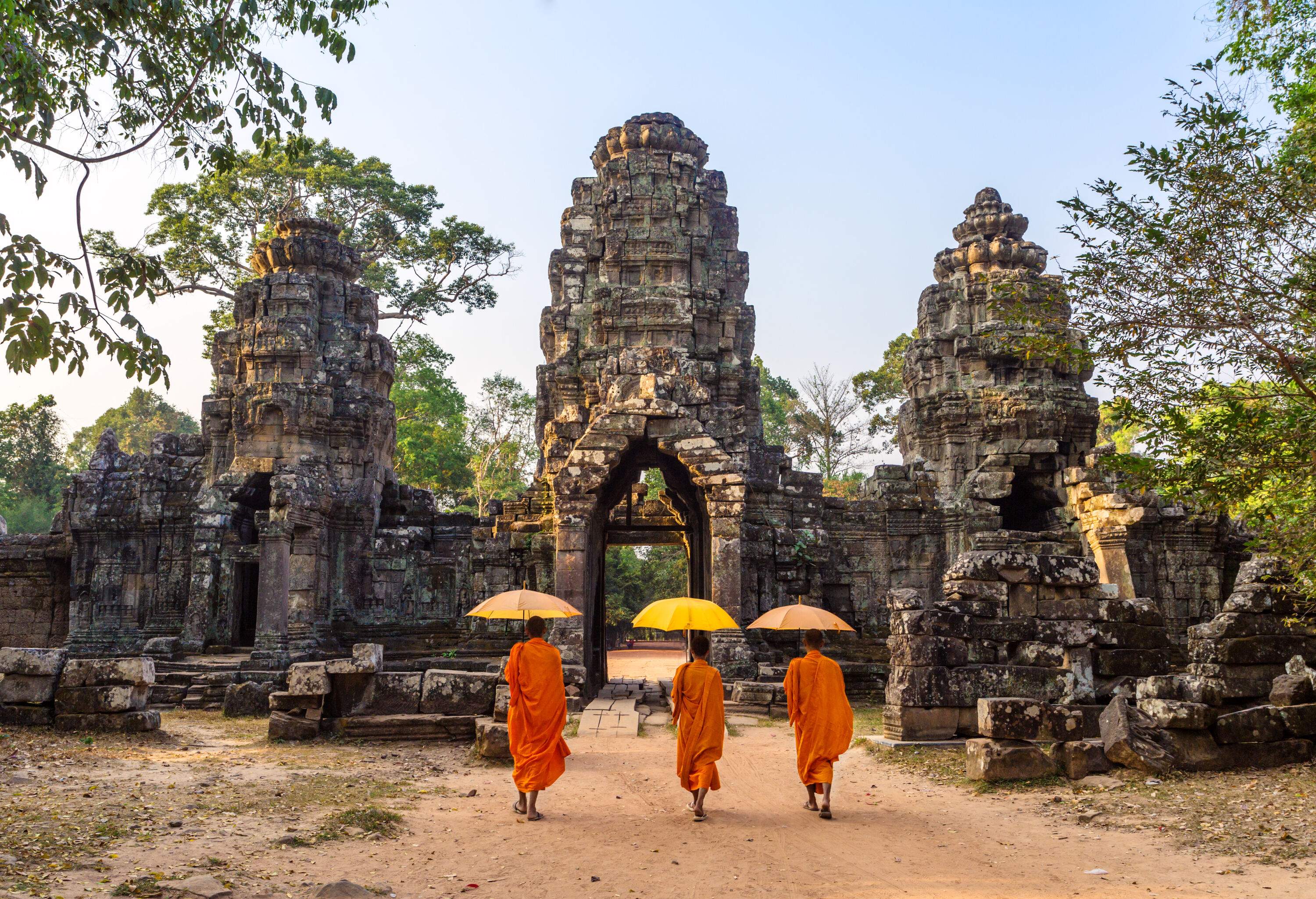 Three Buddhist monks, sheltered by their umbrellas, gracefully traverse the historic stone gate entrance of the majestic Angkor Wat temple complex.