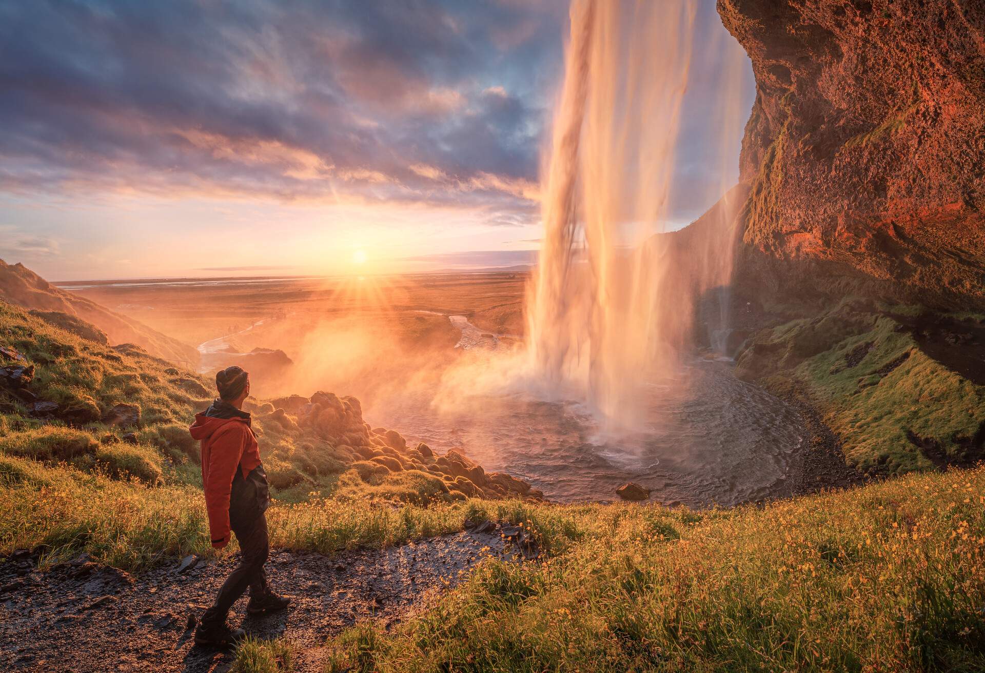 A man stands on a rugged hill with a view of falling waters over a steep rock into a stream on a sunset with dramatic sky.