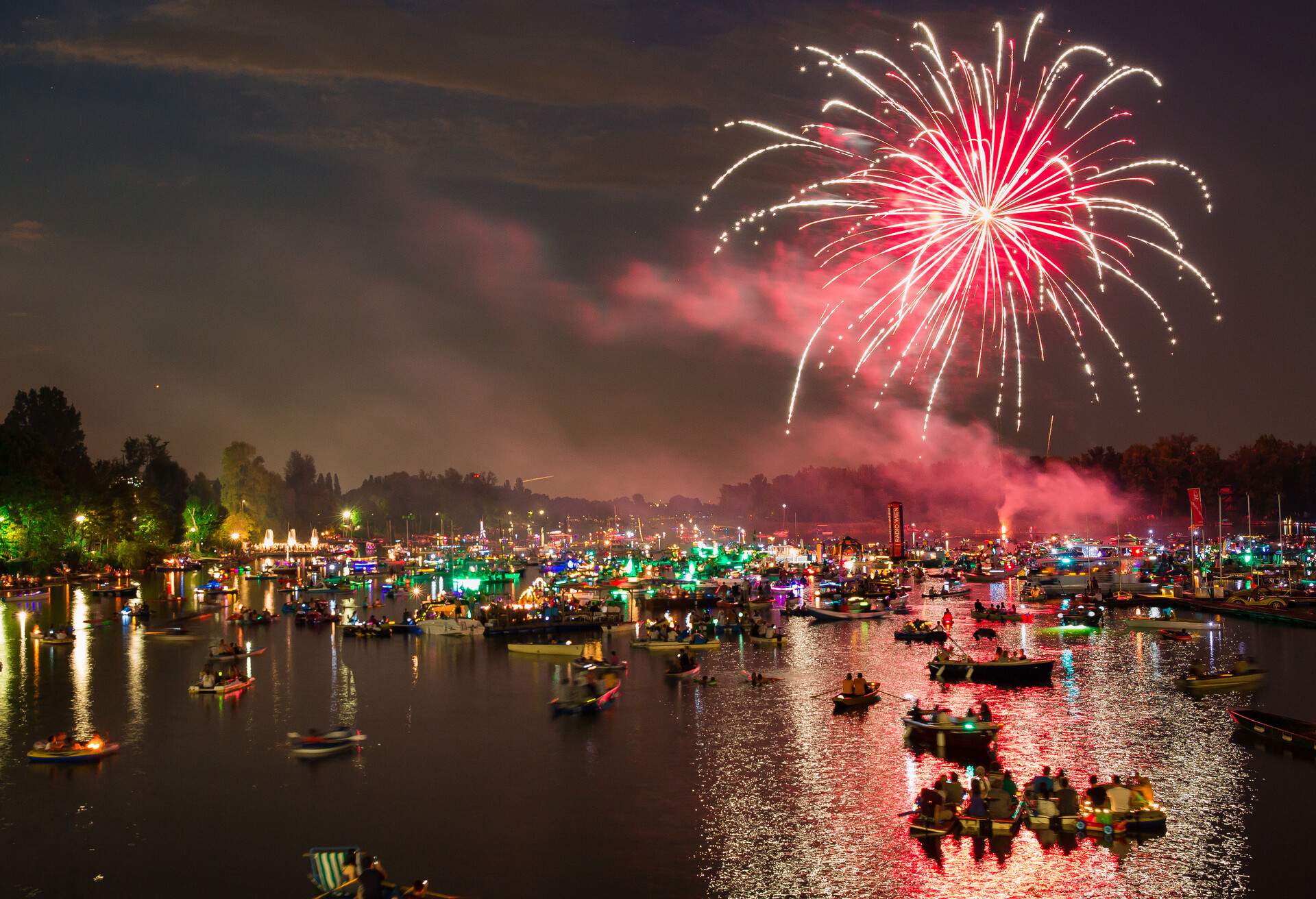 A river crowded with people riding on boats at night, with fireworks lighting up the dark sky.