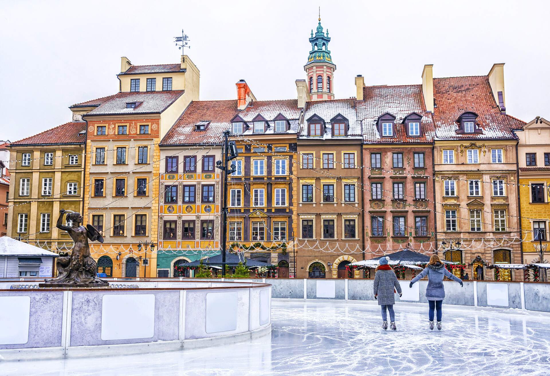 Two girls skate on an ice rink with views of the colourful adjacent Baroque buildings.