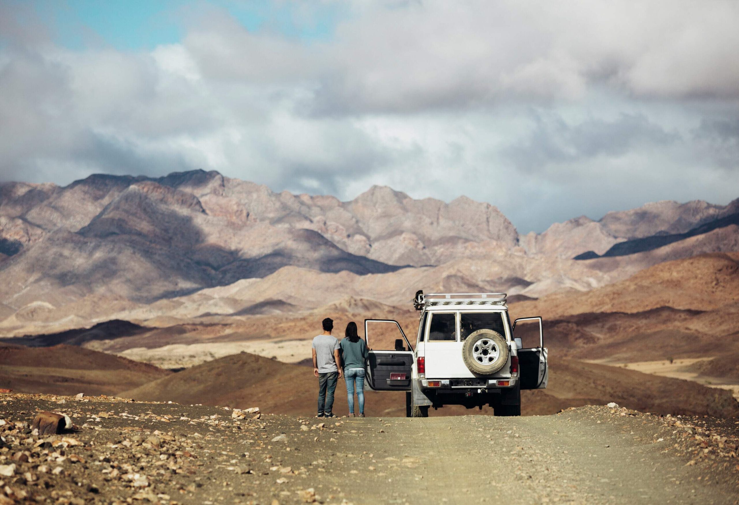 A couple stands next to their vehicle, overlooking the undulating rocky mountain range under the gloomy sky.