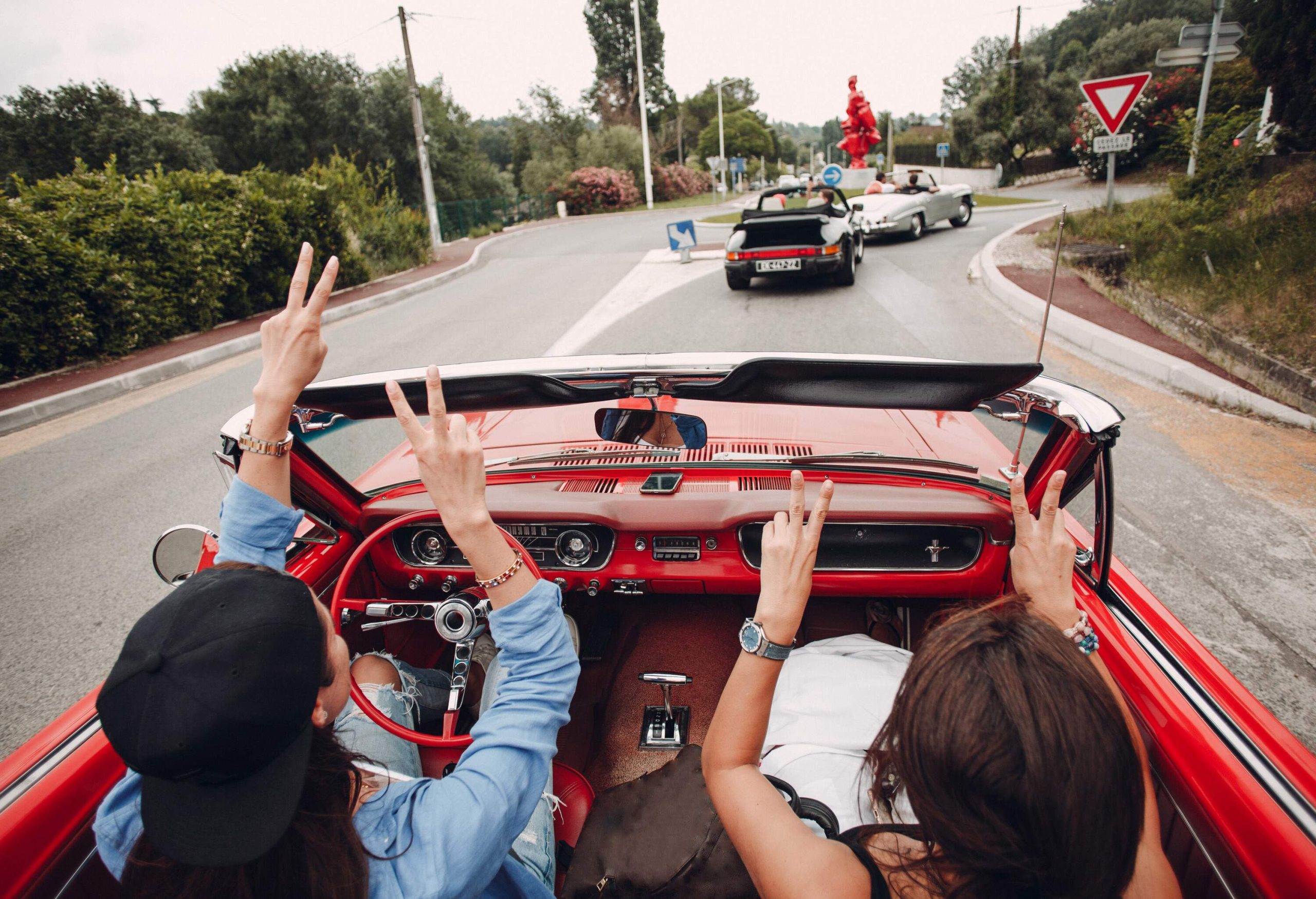 Two women in a red convertible car putting their hands up and making peace signs.