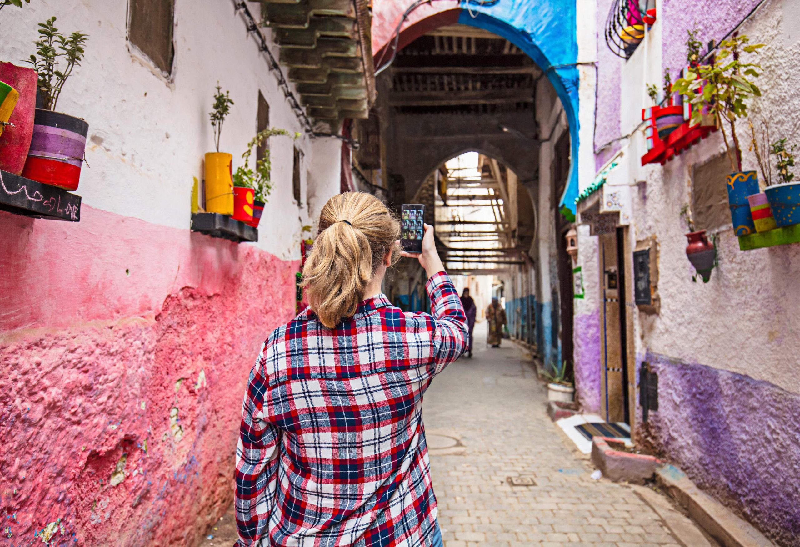 A woman in a chequered shirt uses a phone to take photos of a narrow alley with flower planters on the walls.