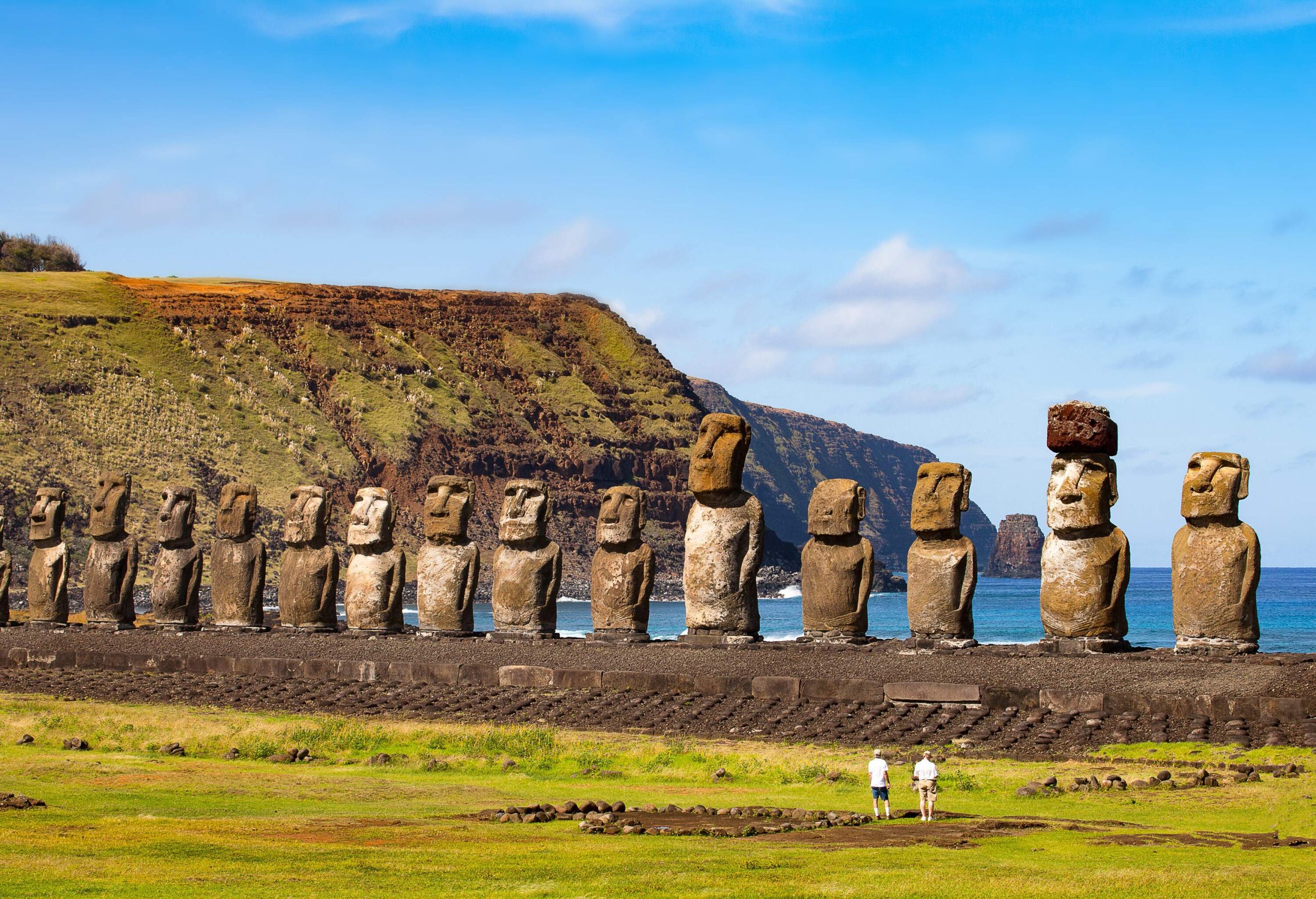 15 majestic Moai statues, gigantic stone sculptures, dominate the scene, with two individuals standing in awe at their bases, while the rugged coastline and a distant coastal cliff provide a dramatic backdrop.
