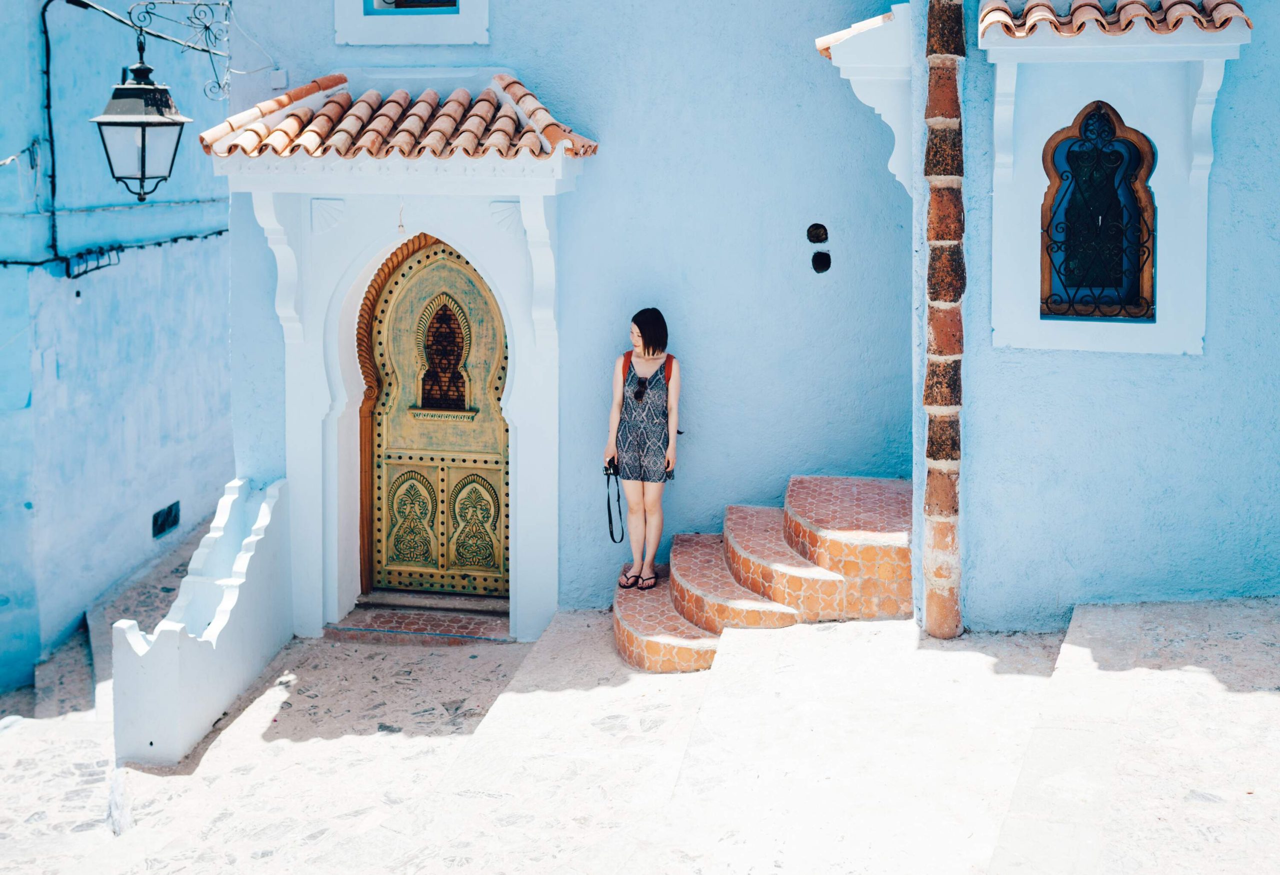 A young lady stands on the stair beside the charming ornate door of a villa with a blue-painted wall.