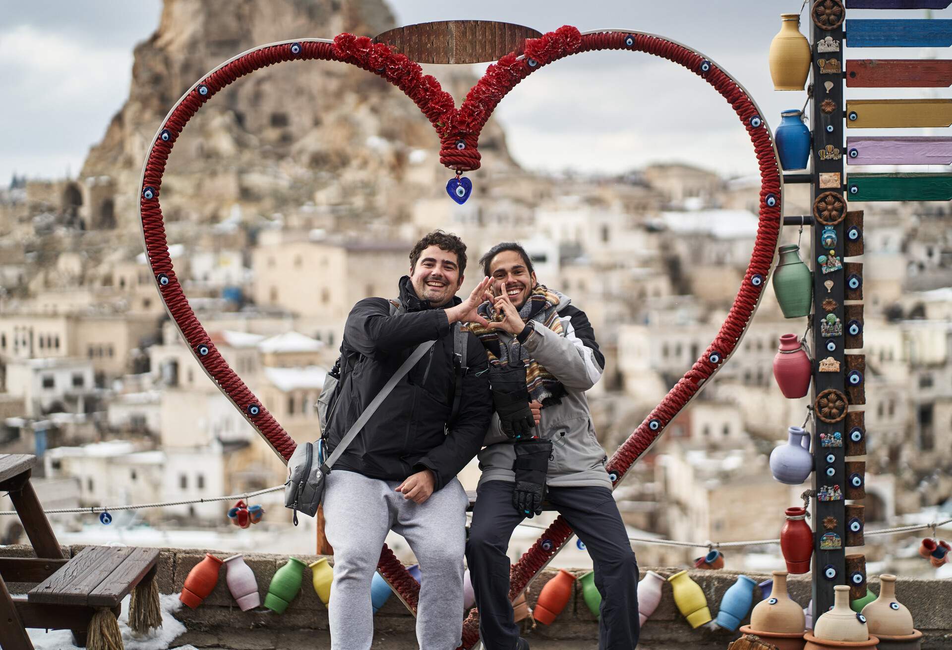 Cheerful boyfriends in outerwear smiling and gesturing heart while sitting on bench near colorful ceramic vases on blurred background of Ortahisar castle in Cappadocia, Turkey