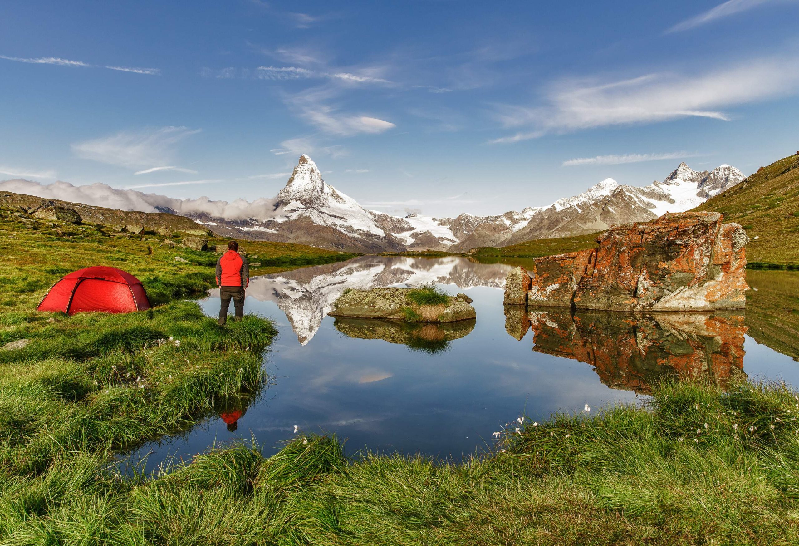 A camper is admiring the stunning view of the Swiss Alps while standing next to a dome tent beside a lake.