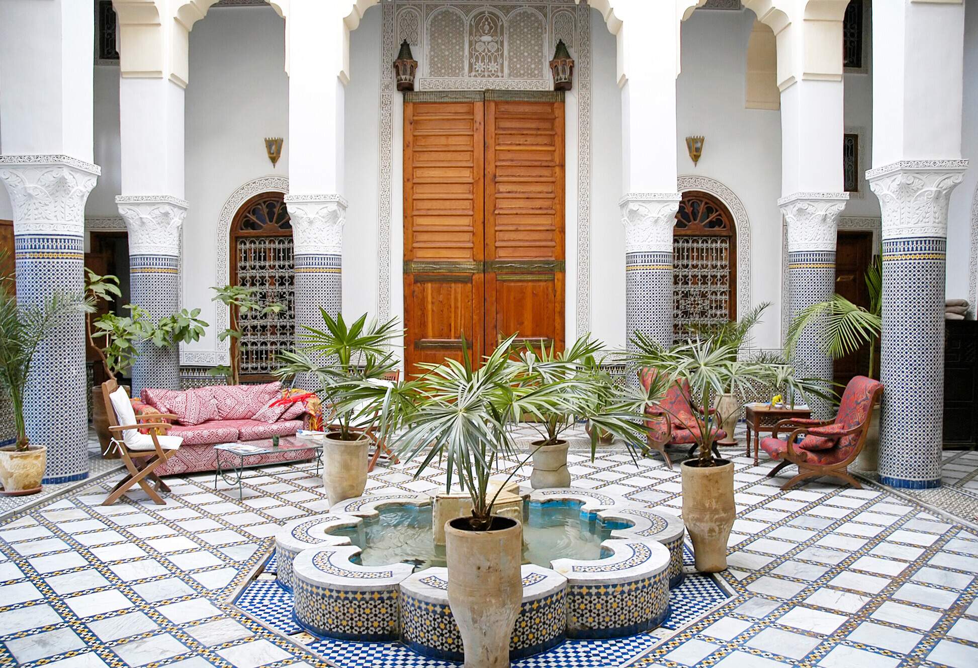 A traditional Moroccan house unfolds around a serene courtyard, where a fountain takes centre stage, surrounded by lush planters and graceful columns.