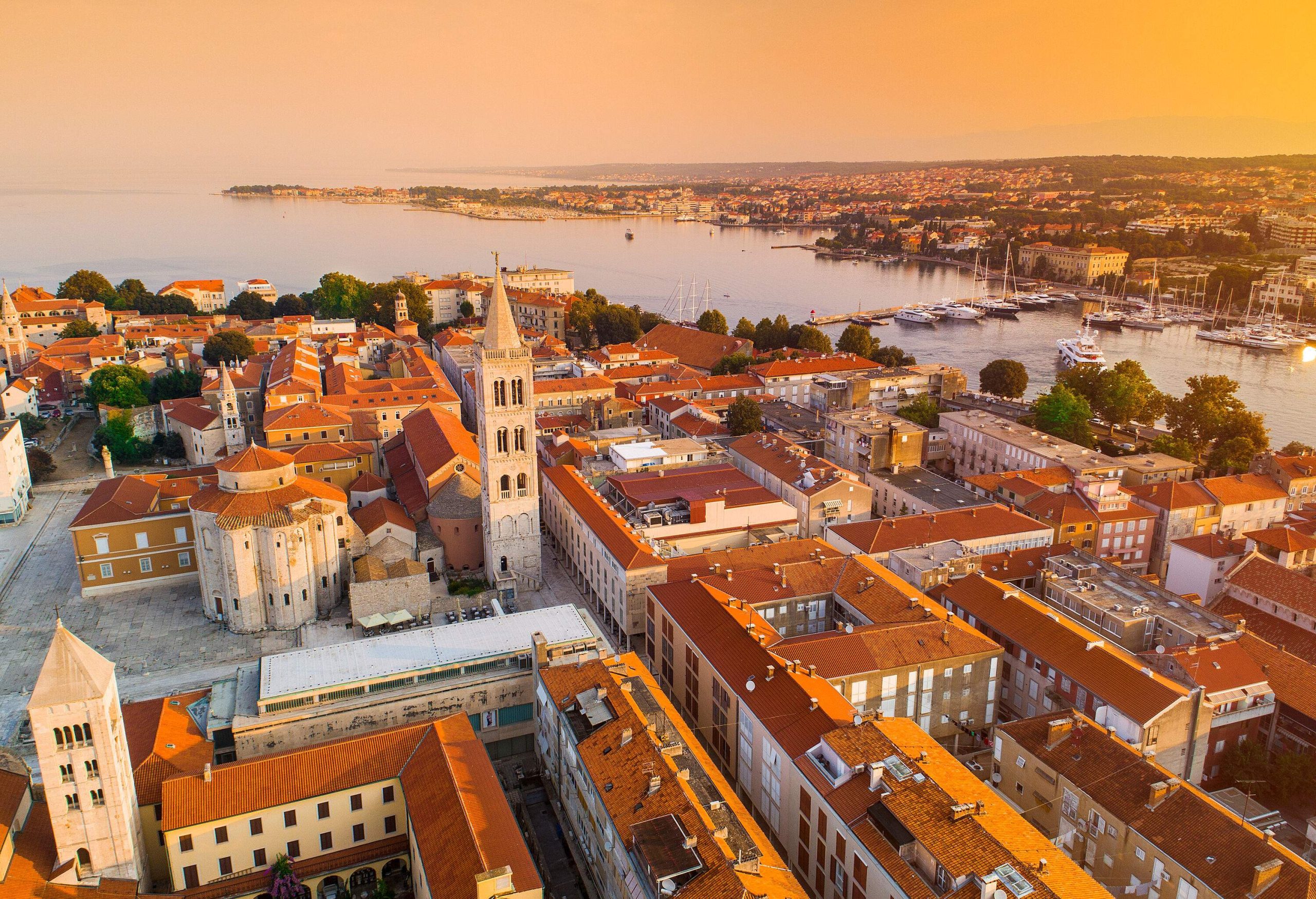 A church's pointed bell tower rises above a cluster of buildings along a harbour with anchored boats under the orange sky.