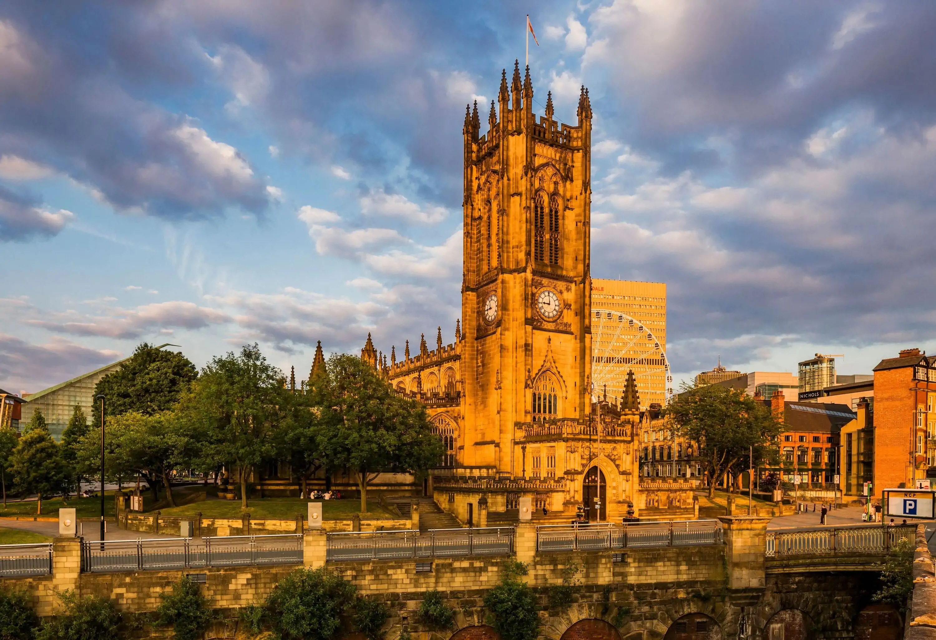 A street along the sunlit Manchester Cathedral, a church with a square clock tower and sharp pinnacles.