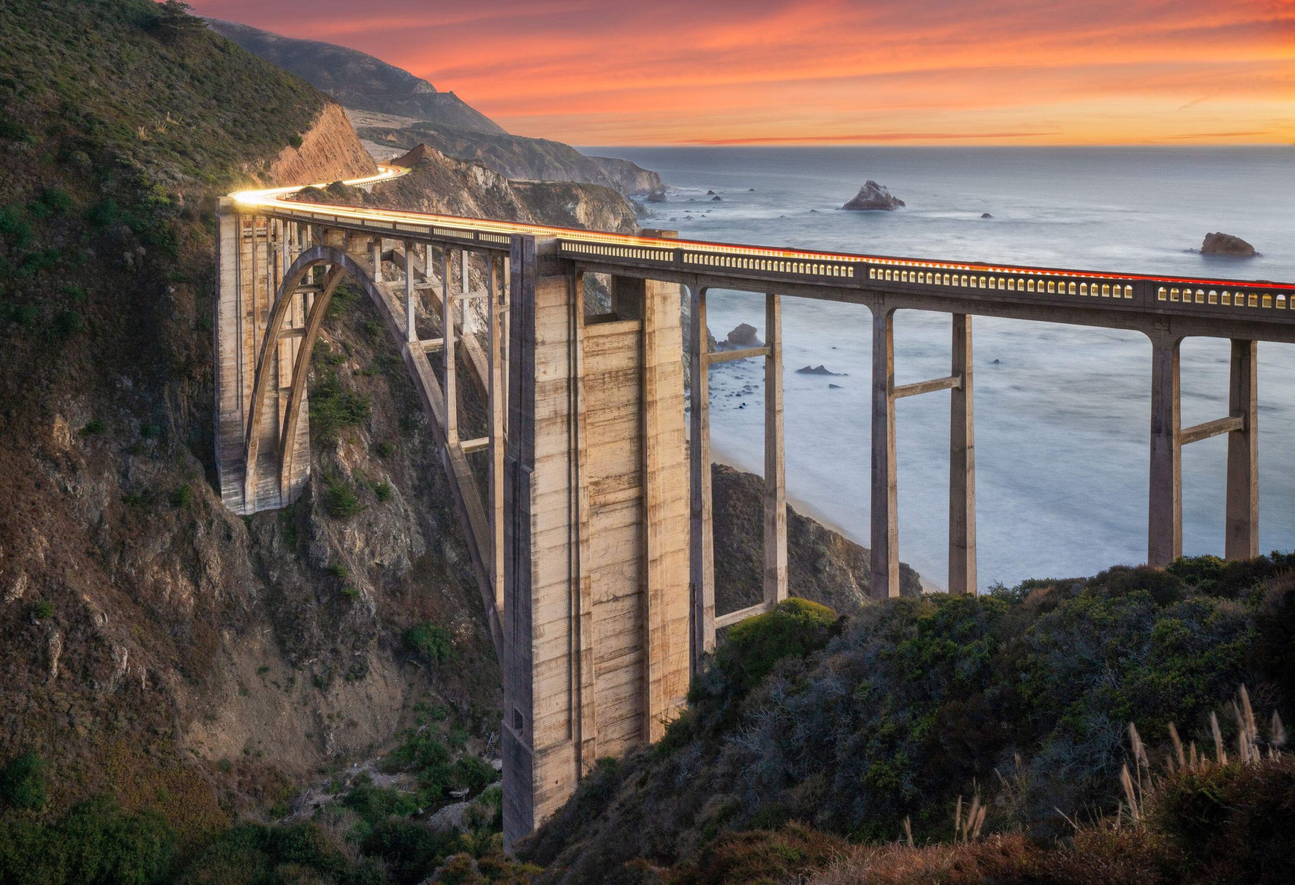 A tall, open-spandrel arch bridge between steep and crumbling cliffs on the coastline on a dramatic sunset.