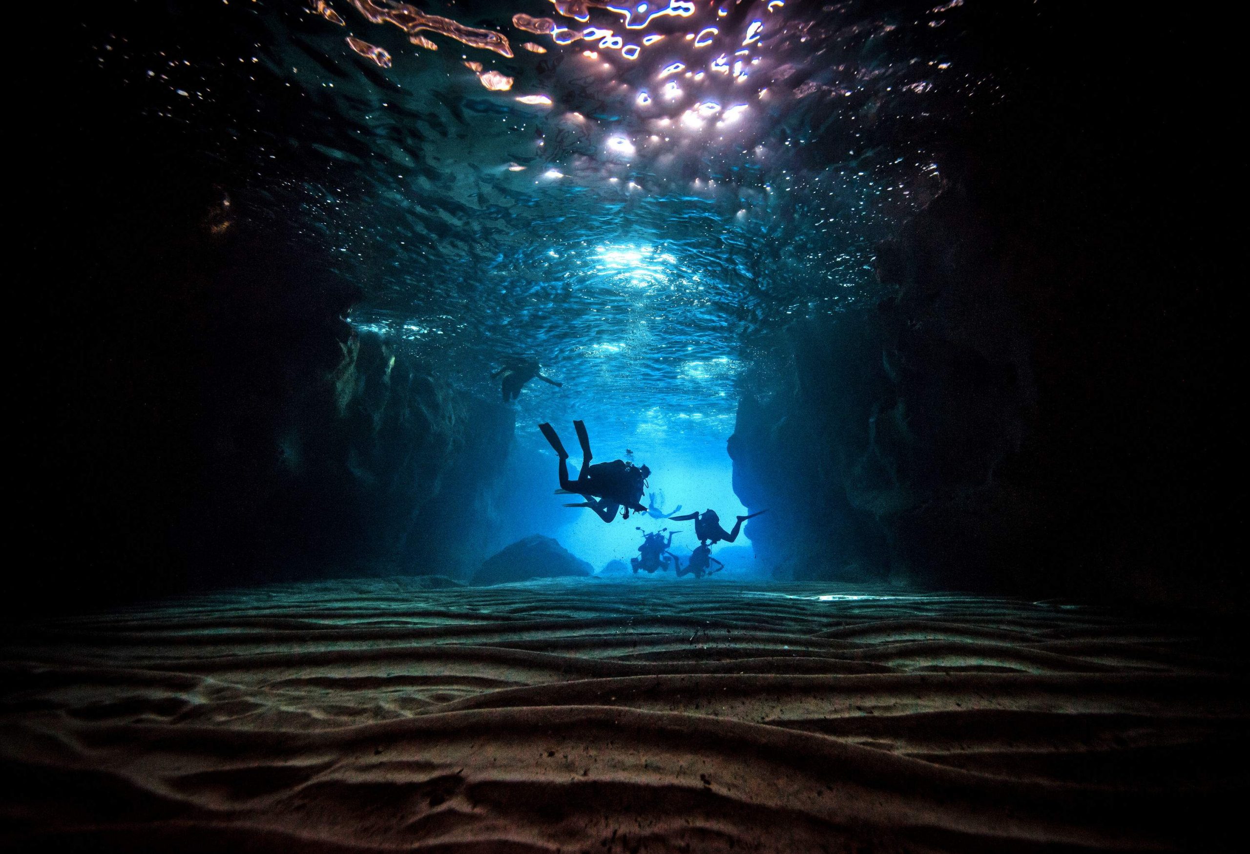 A group of scuba divers swimming across a dimly-lit underwater cave.