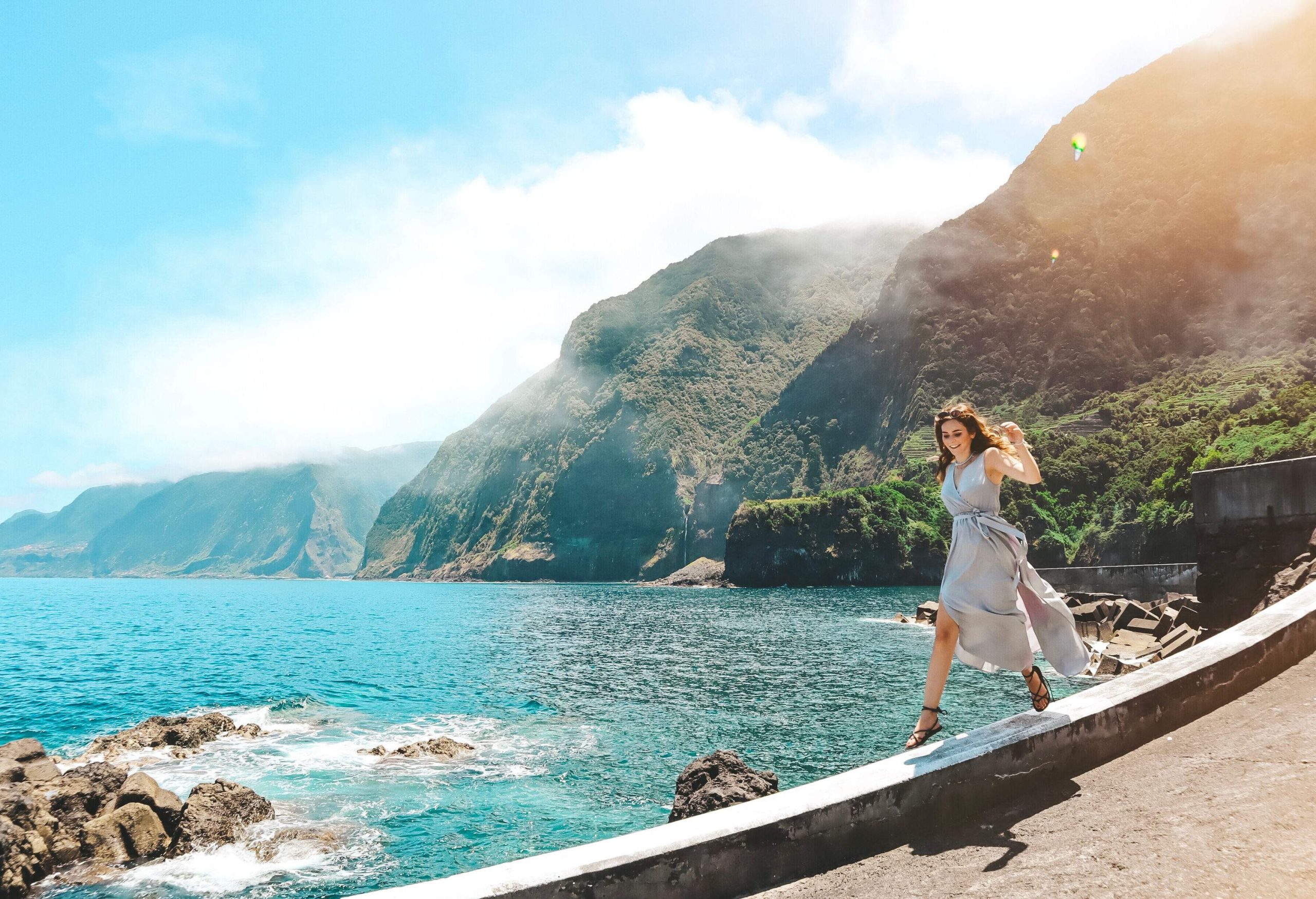 A woman striding cheerfully on a ledge by the sea with mountains in the background.