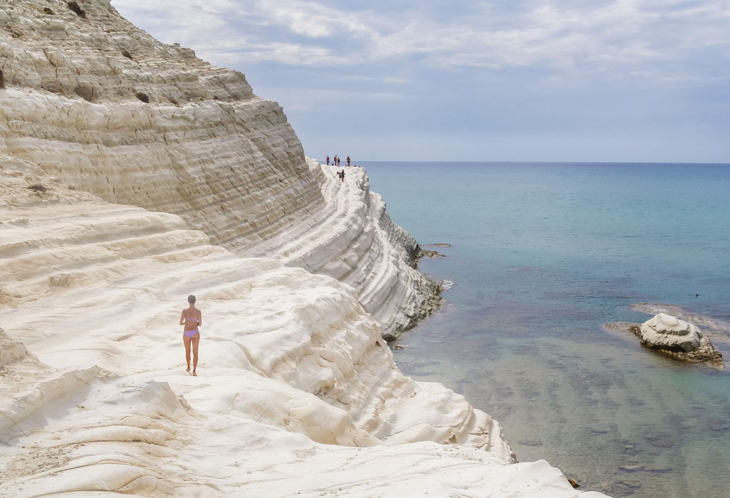 A woman in a bikini walking along a tiered white cliff by the sea.