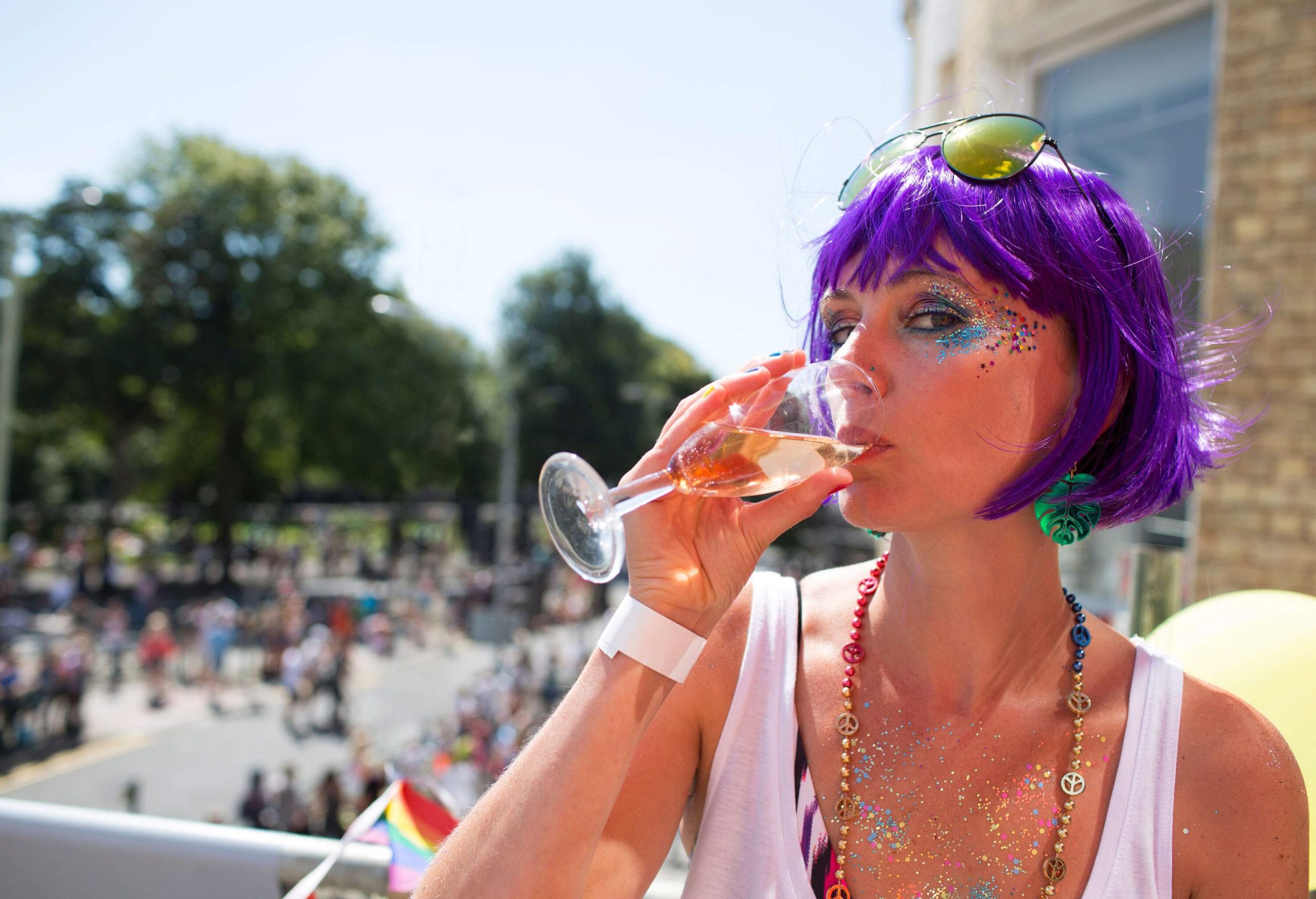 A young woman wearing a purple wig and sunglasses sipping champagne outdoors on a sunny day.