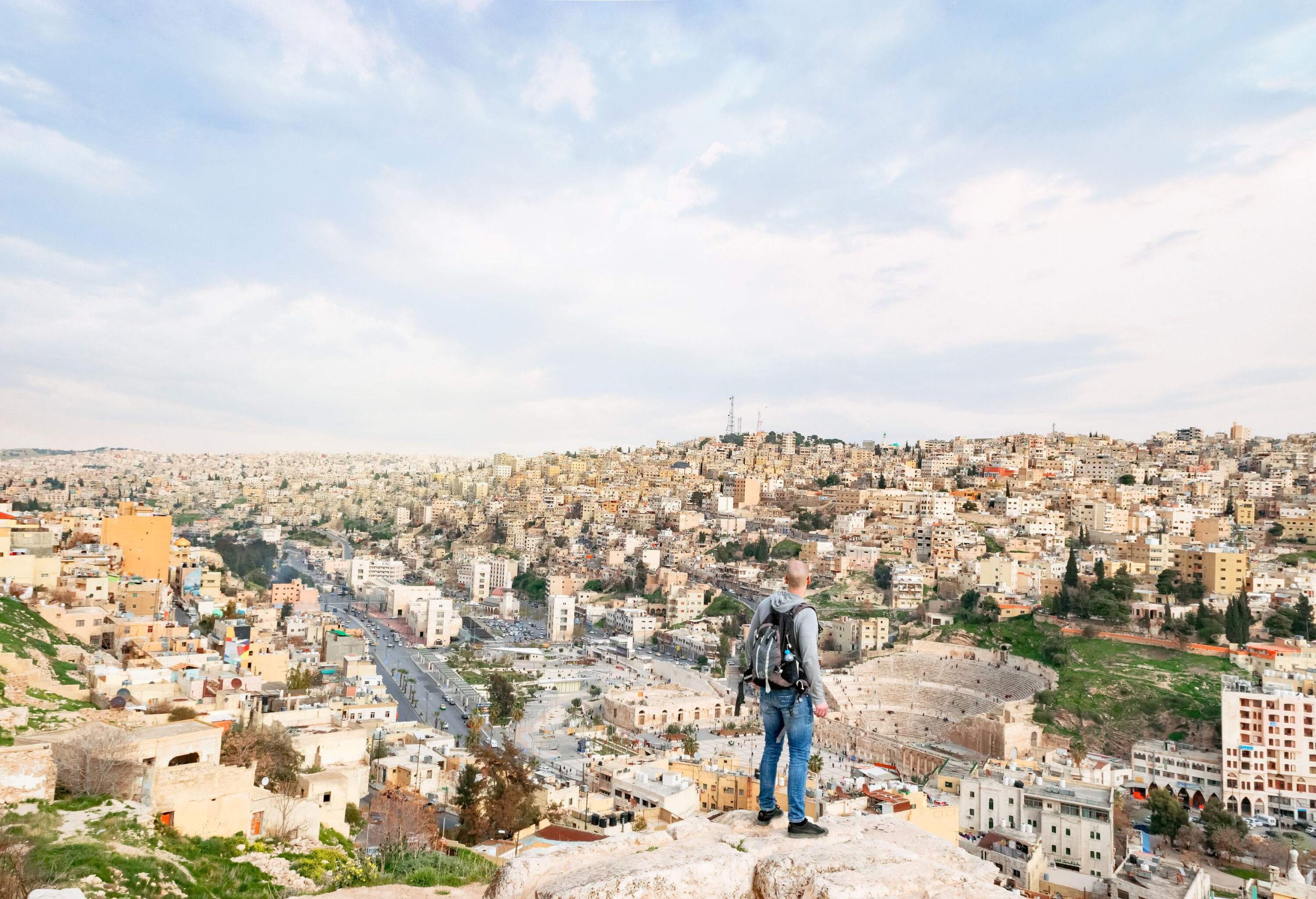 A man stands atop a hill overlooking a bustling metropolitan with a cluster of buildings uphill and a historic Roman amphitheatre.