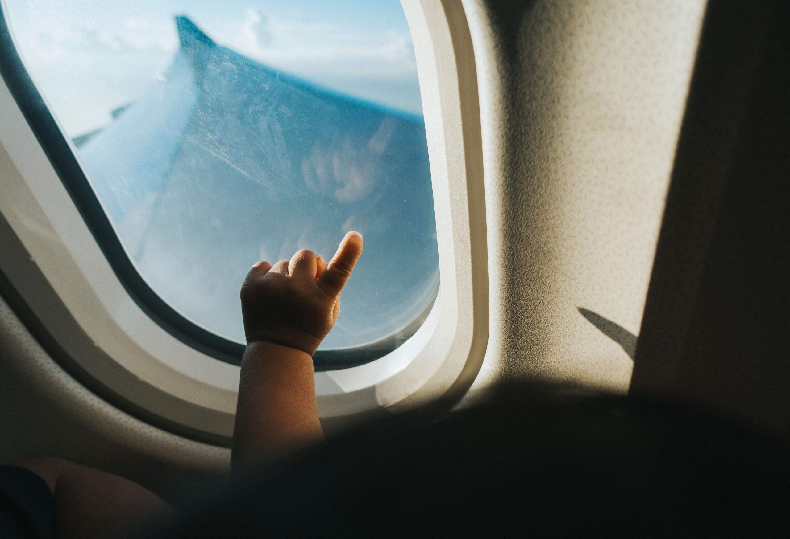 A toddler points at an aeroplane window with a view of a cloudy blue sky.
